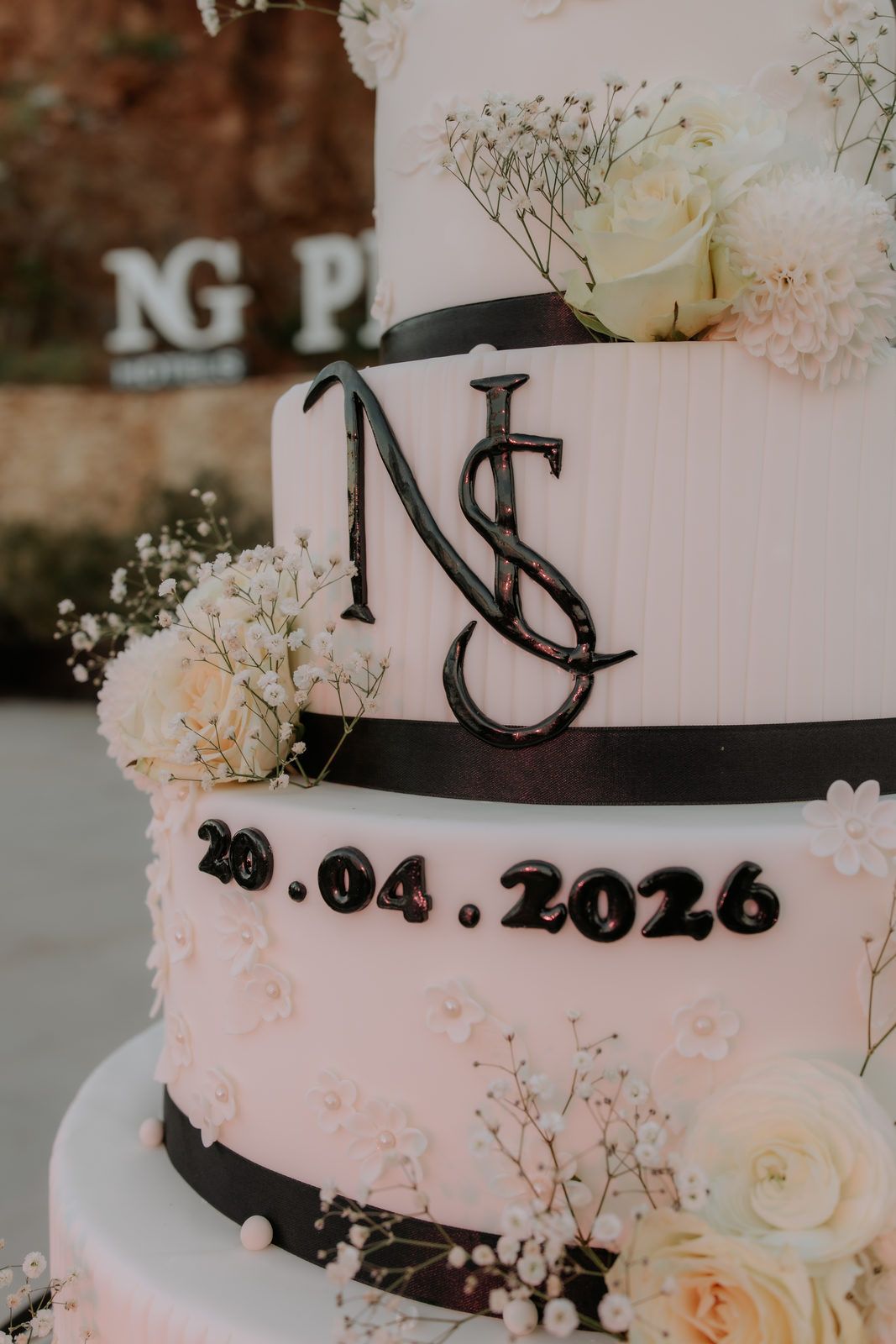 Elegant three-tier white wedding cake with black ribbon, monogram topper, date 20.04.2026, and white floral decorations.