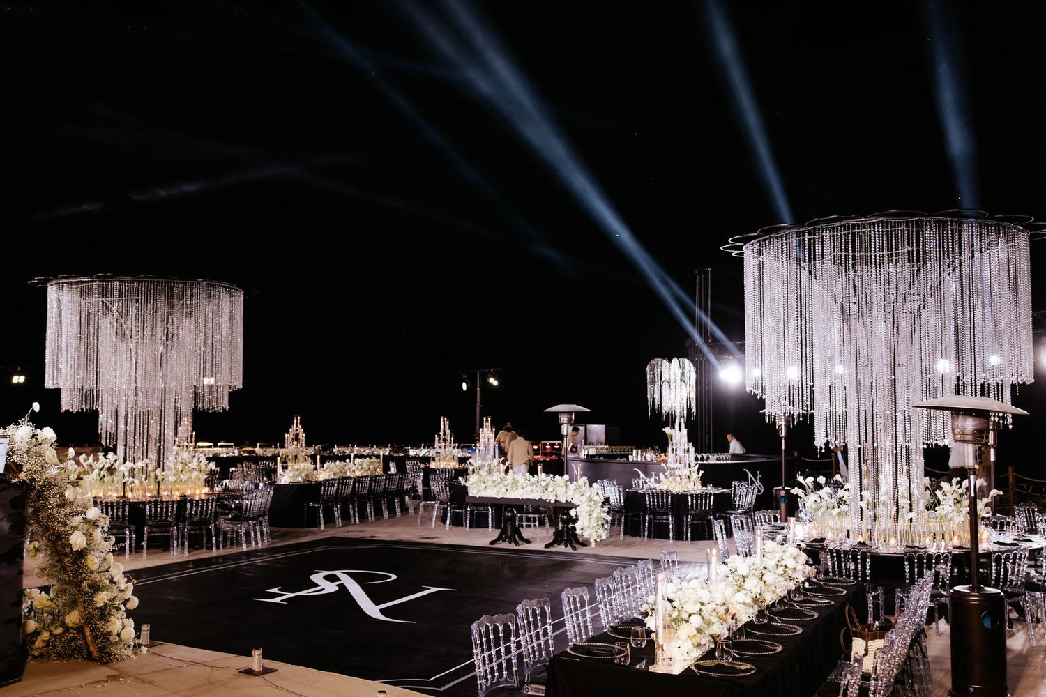 Elegant outdoor wedding reception with crystal chandeliers, white floral arrangements, and dramatic light beams at night.