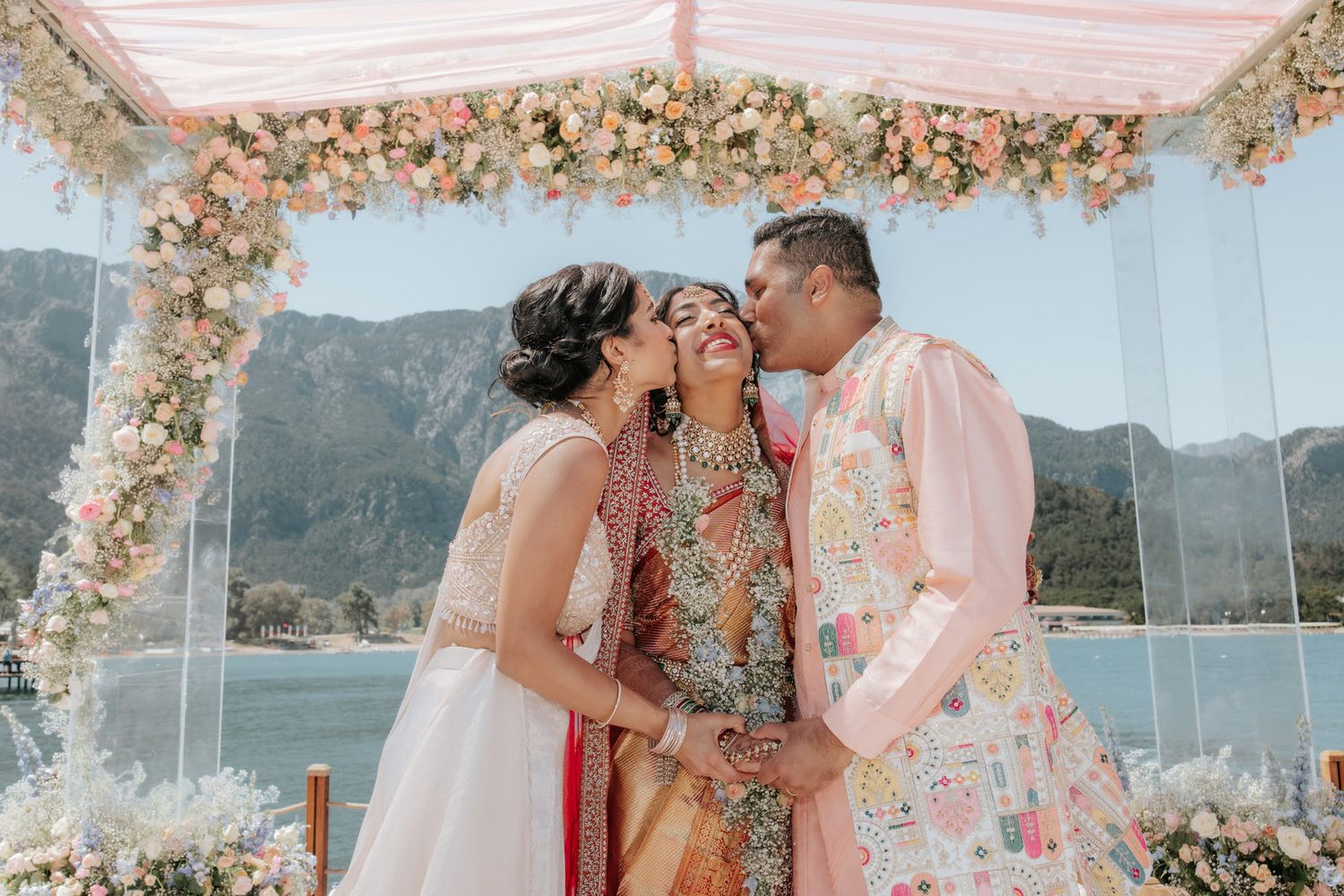 Bride in colorful lehenga receives kisses from parents under floral mandap with mountain lake backdrop.