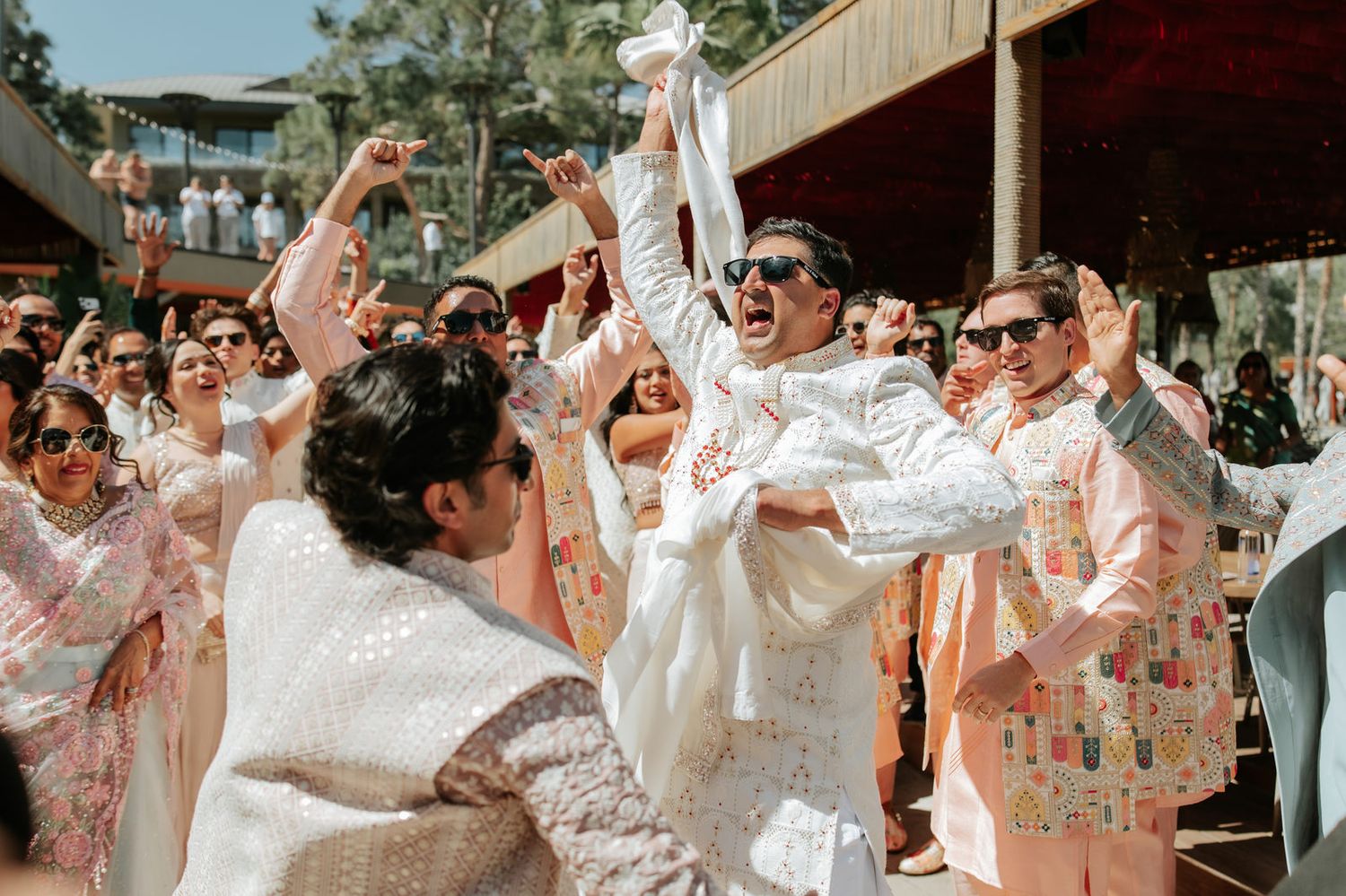 Groom in white sherwani celebrates joyfully with guests during an Indian wedding baraat.