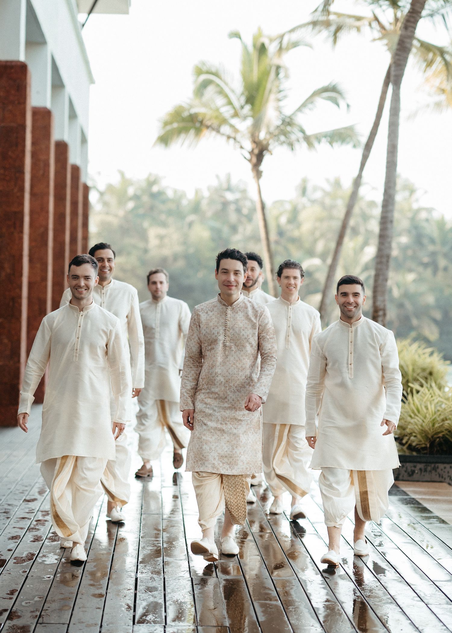 A groom in ornate kurta leads groomsmen in cream sherwanis walking outdoors at an Indian wedding.