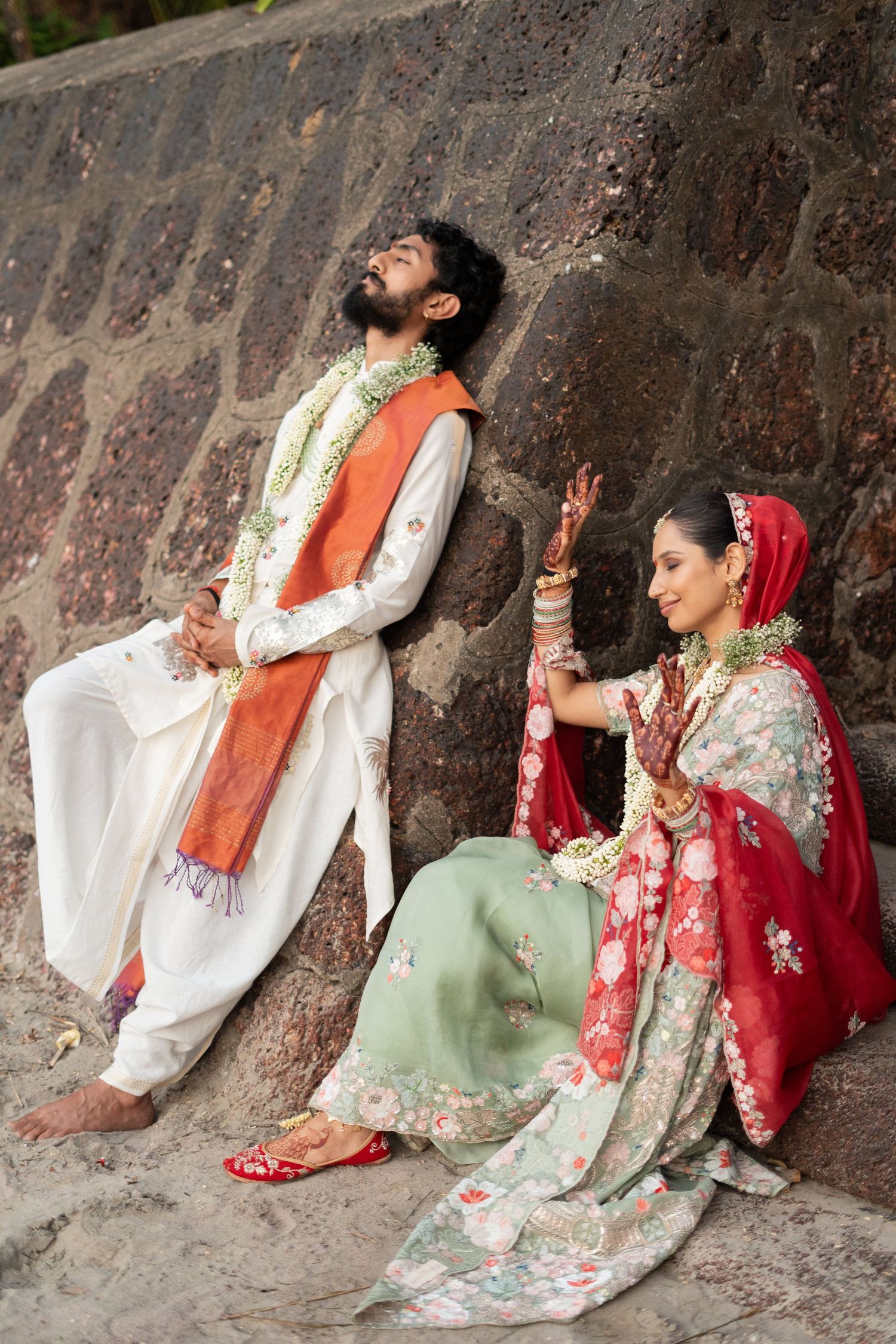 Indian bride and groom in traditional wedding attire sitting against a stone wall, adorned with floral garlands.