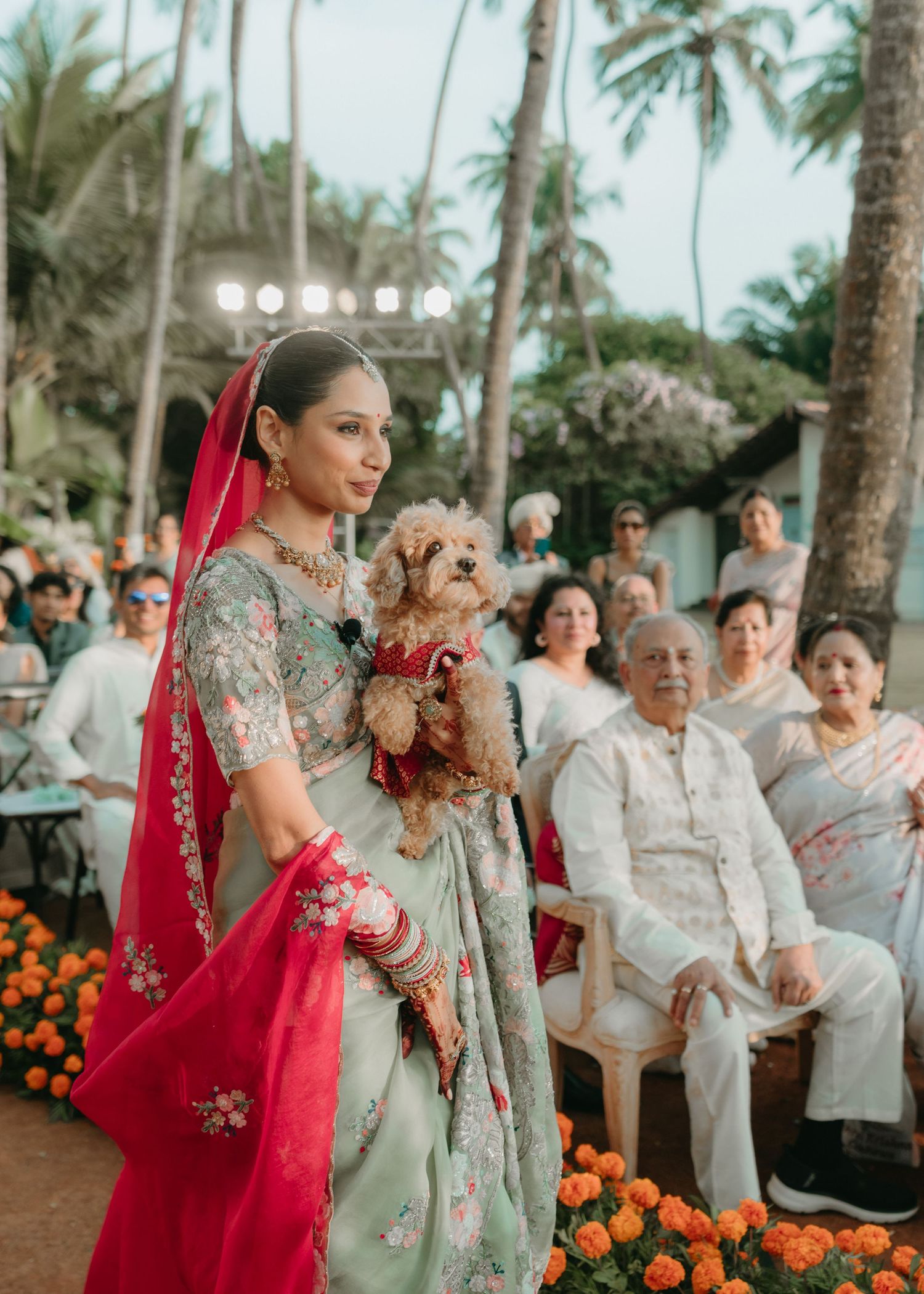 Bride in sage green lehenga with red dupatta holds a dog at an outdoor Indian wedding ceremony.