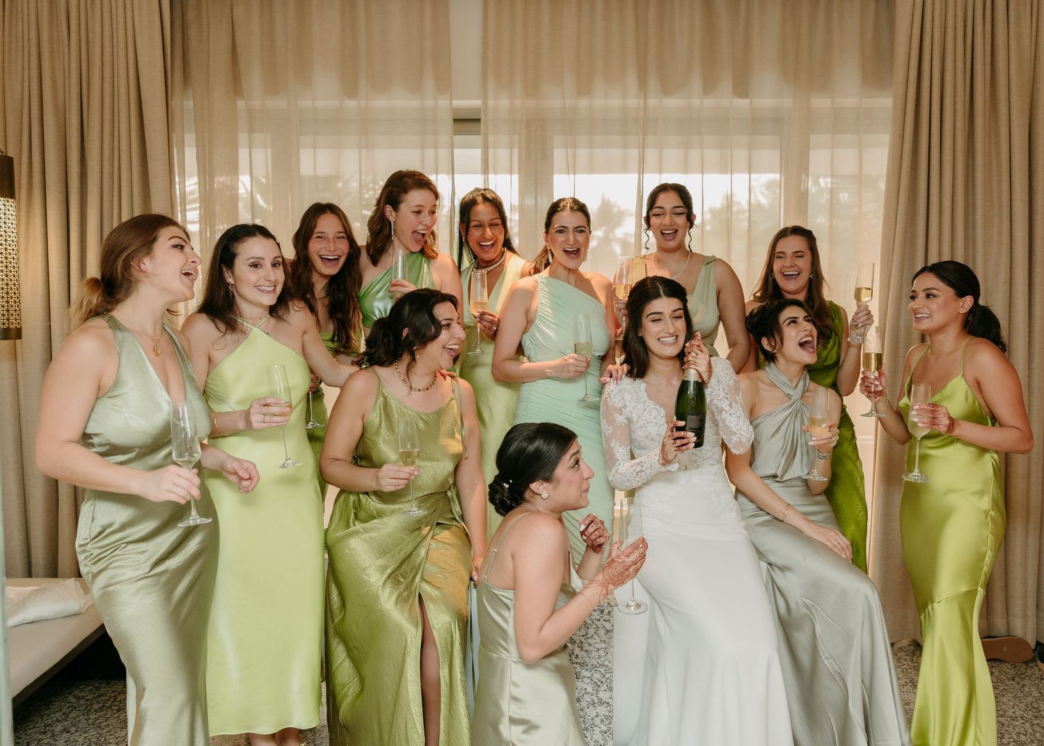 Wedding party wearing coordinated mint green and sage silk dresses laugh together in a brightly lit room.