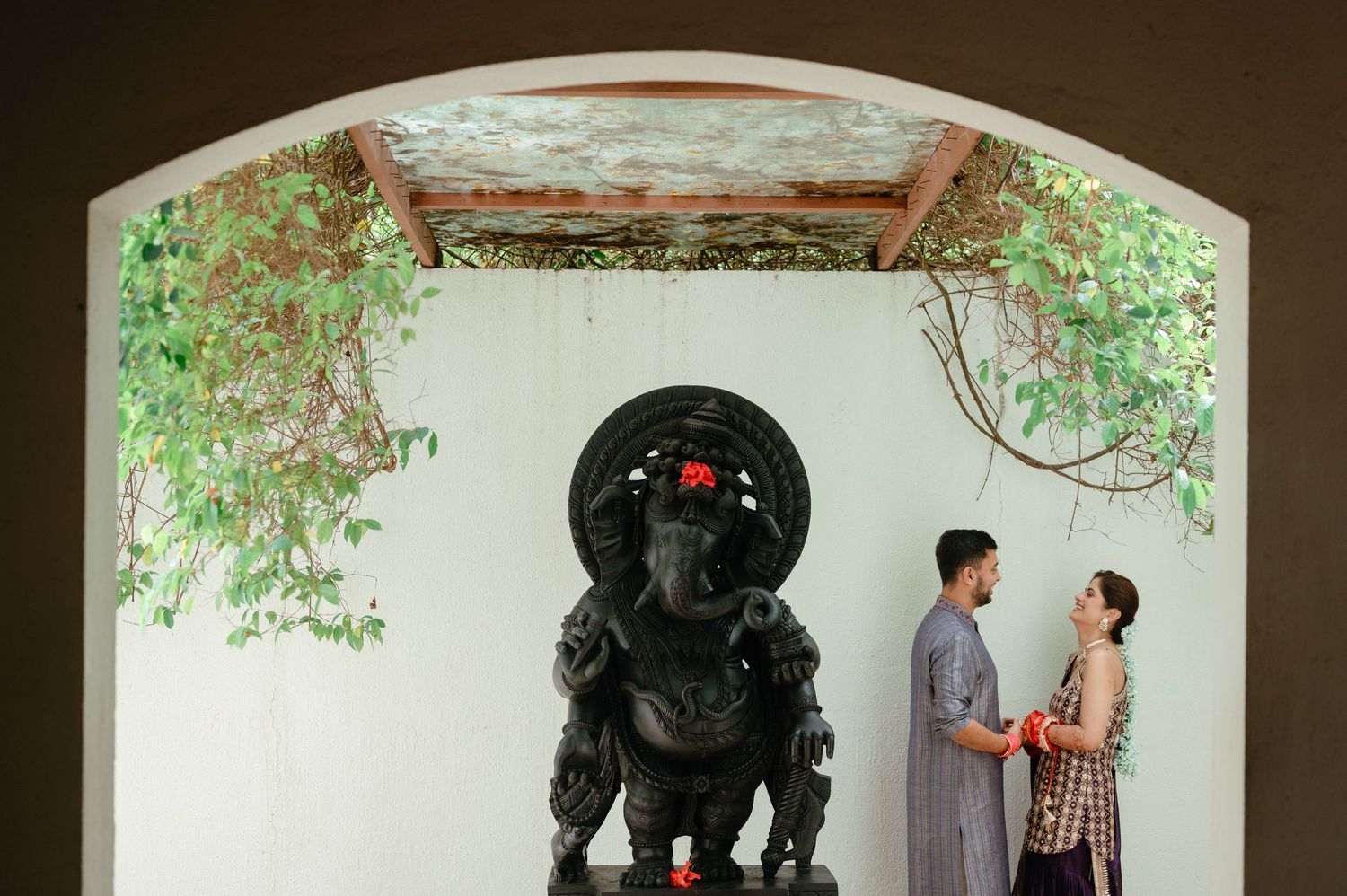Couple standing beside a large Ganesha statue in an arched alcove with lush green vines overhead.