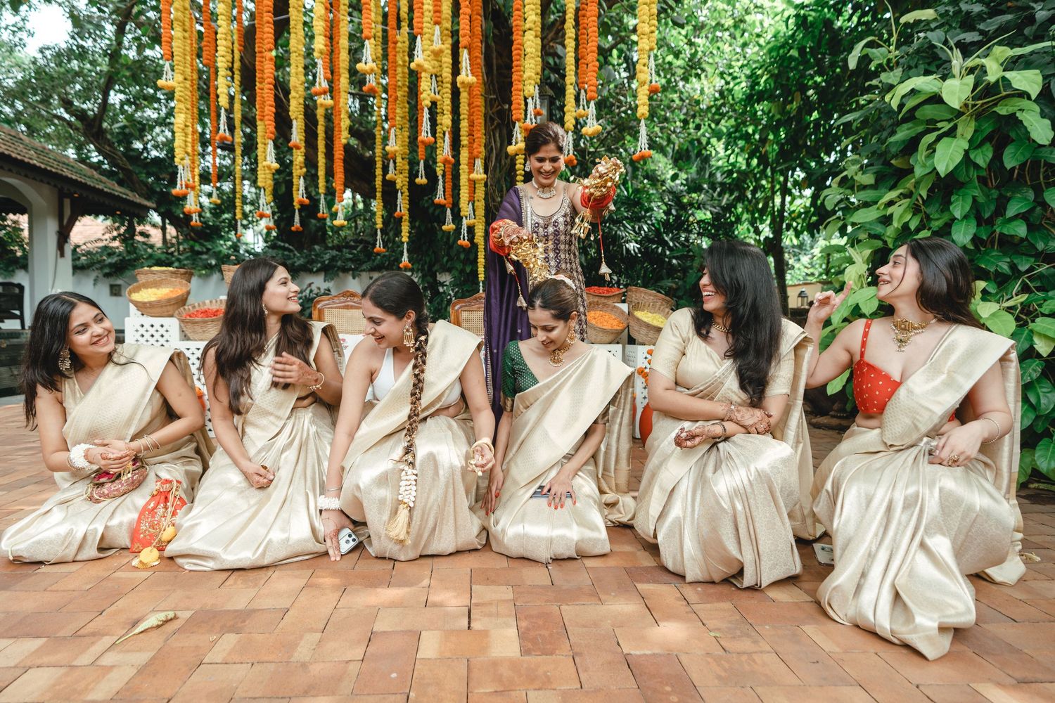 Women in gold sarees sit together laughing at an Indian celebration with marigold flower decorations overhead.