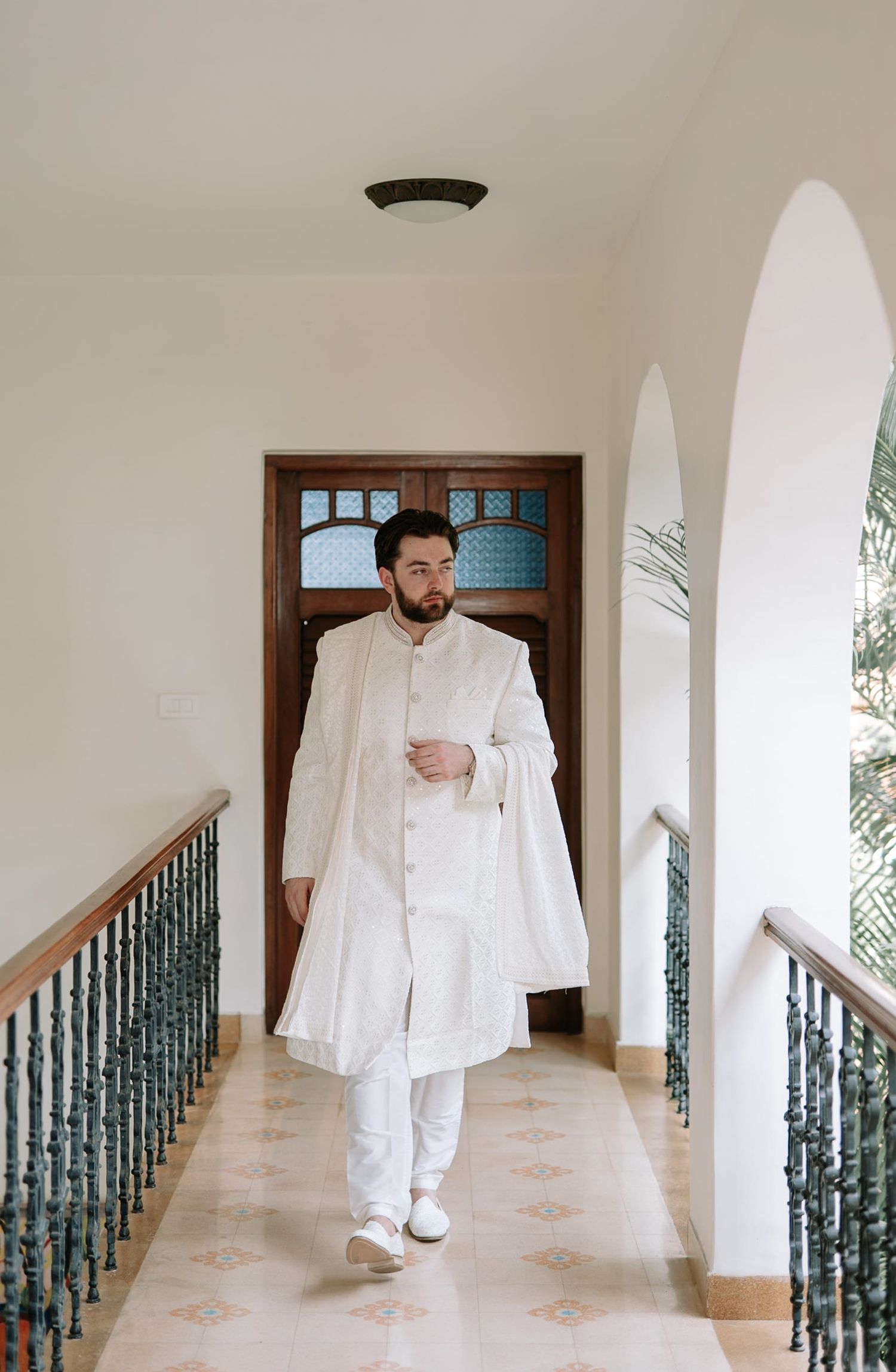 A groom in a white sherwani walks down an elegant hallway with arched doorways and ornate railings.