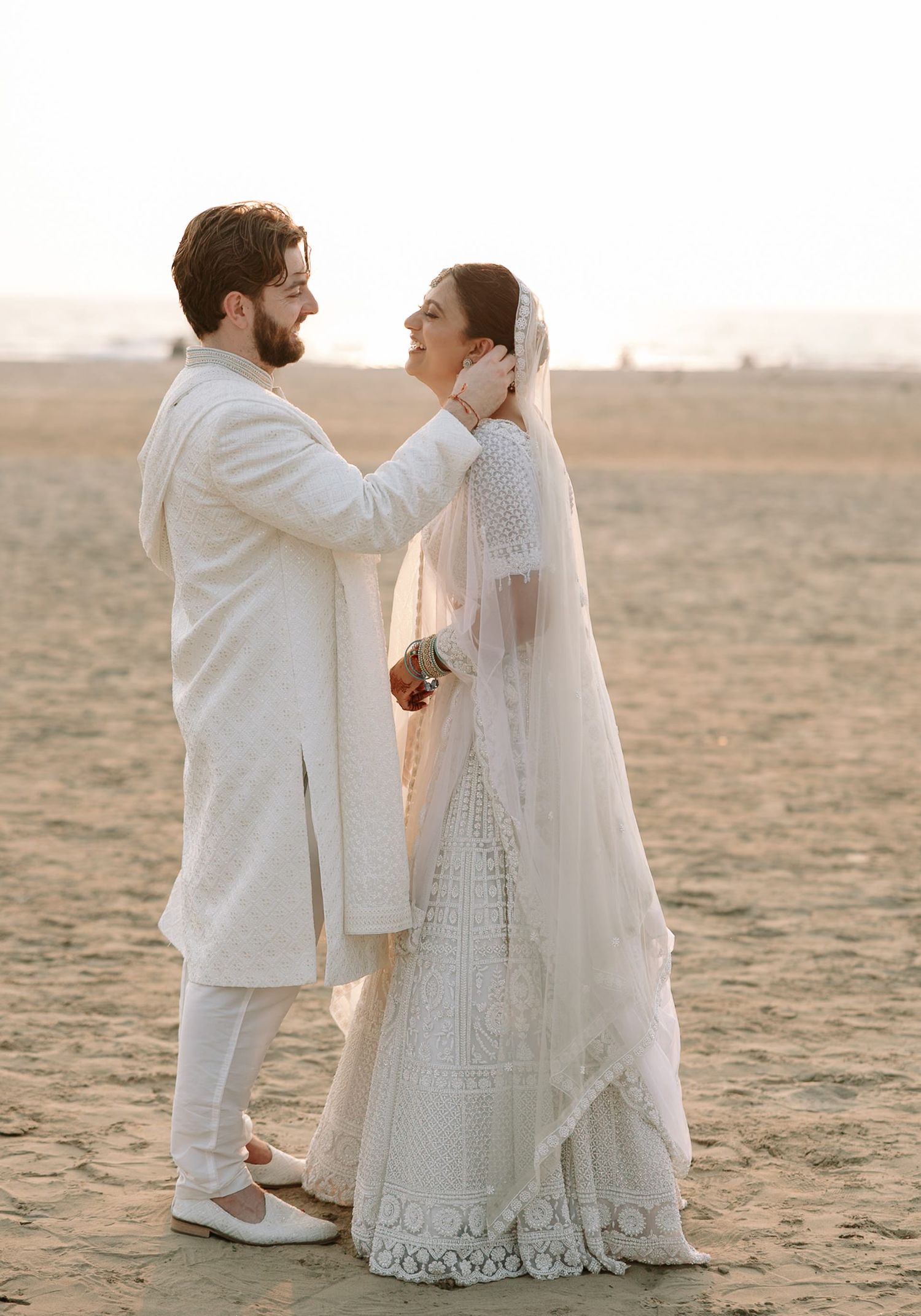Couple in white wedding attire on sandy beach, groom in sherwani adjusting bride's veil at golden hour.
