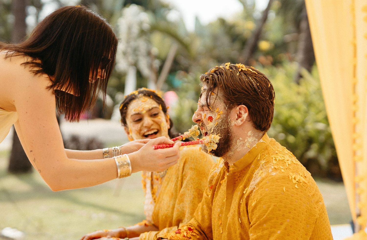 Couple in yellow outfits celebrating Haldi ceremony outdoors, laughing as turmeric paste is applied.
