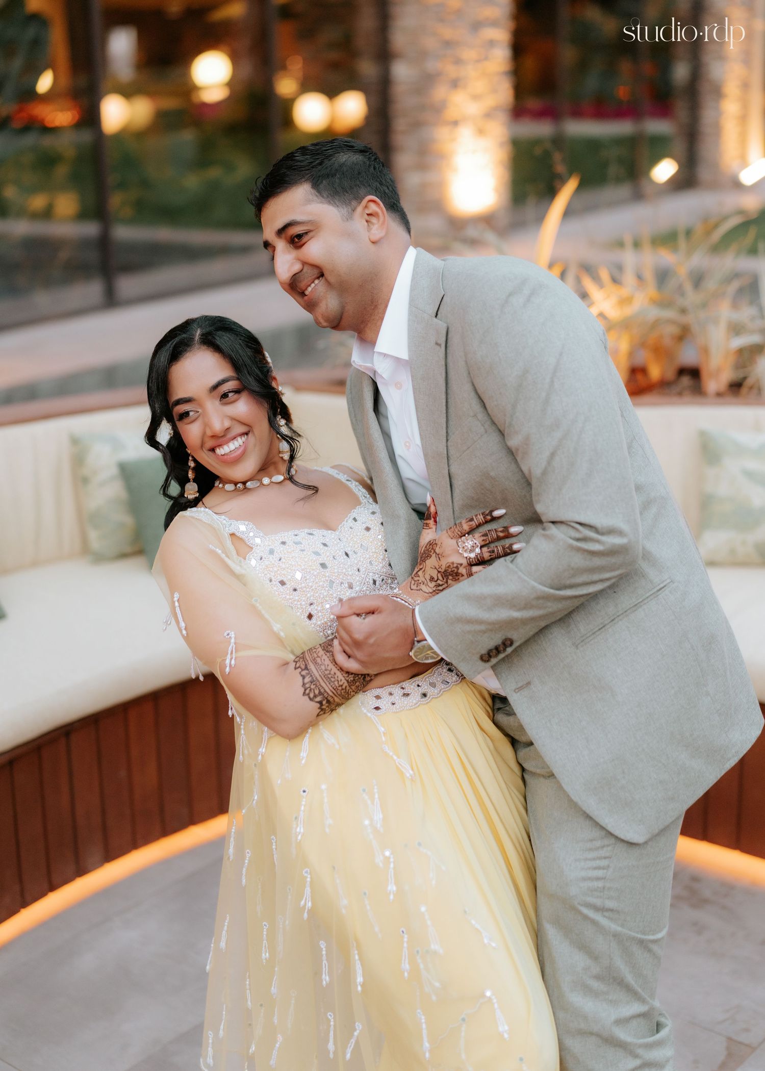 Indian couple in yellow lehenga and grey suit smile joyfully together during elegant outdoor wedding celebration.