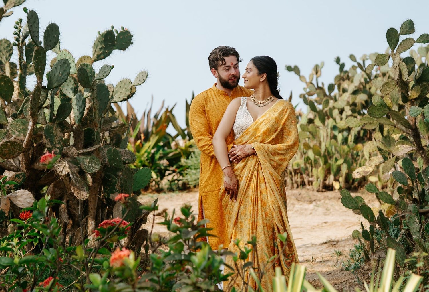 Couple in matching yellow traditional Indian outfits embracing in a lush cactus garden on a sunny day.