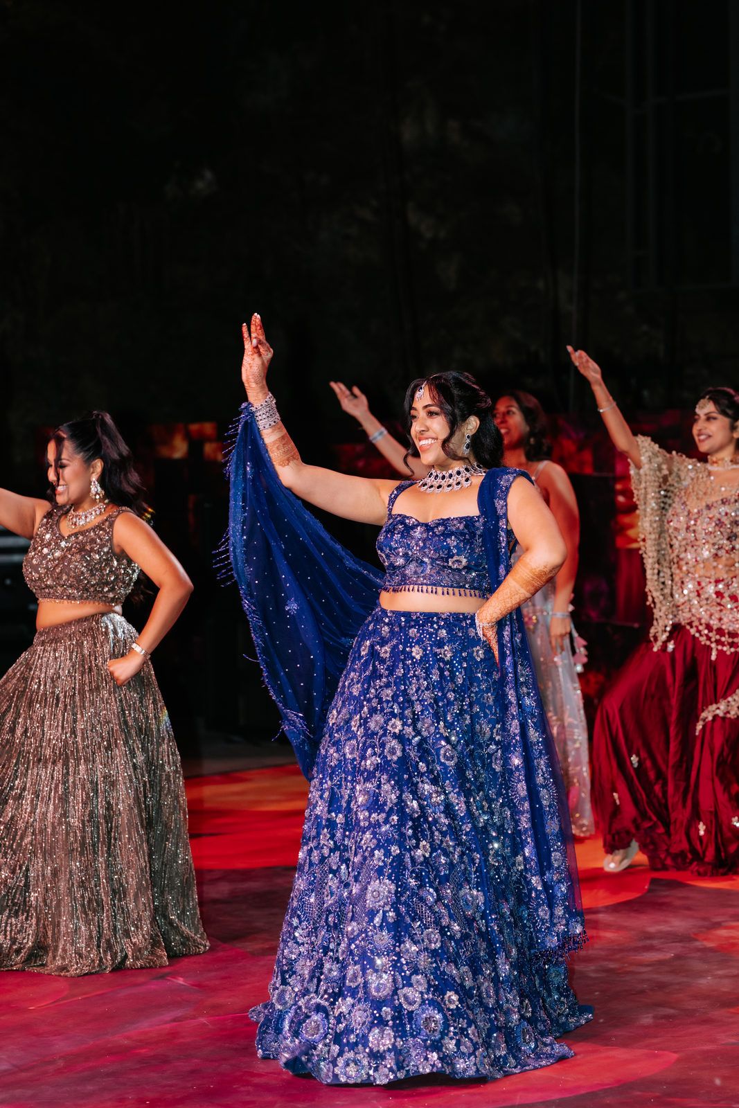 Women dancing in vibrant Indian attire, featuring a blue embroidered lehenga at a festive celebration.