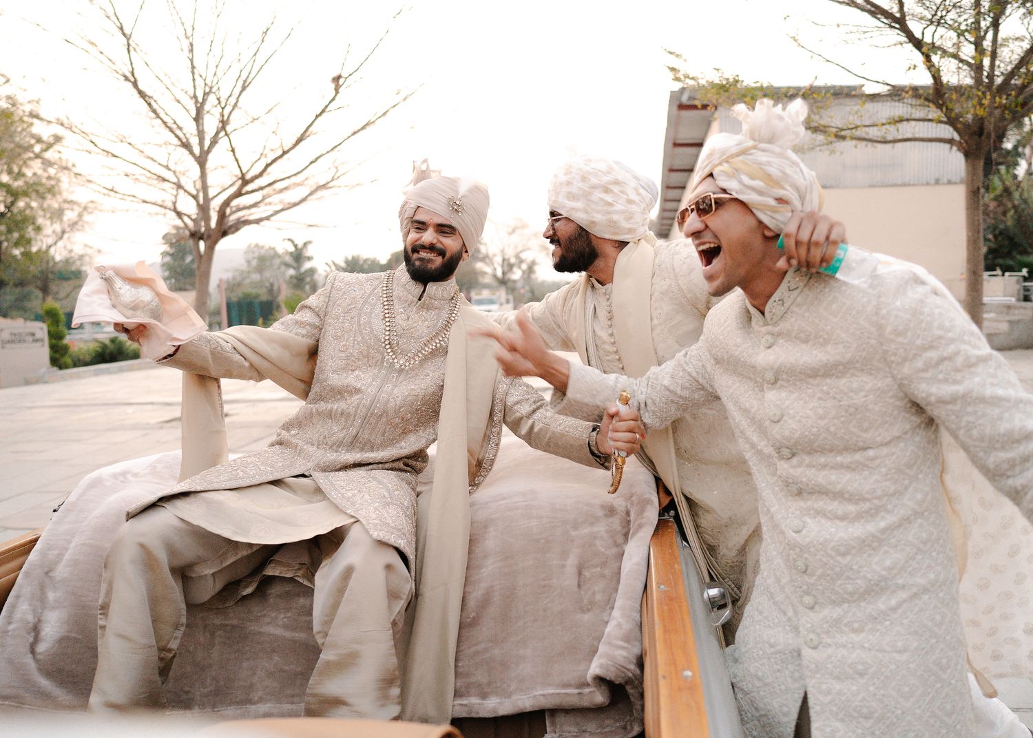 Three men in traditional Indian sherwani and turban laughing joyfully during a wedding celebration outdoors.
