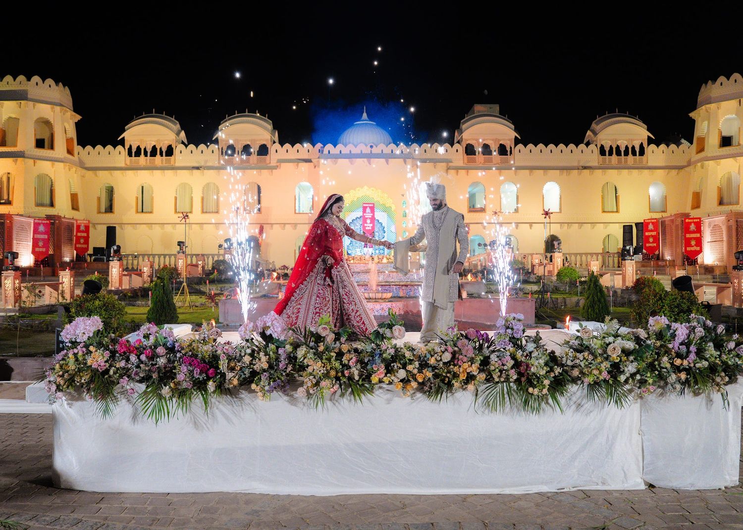 Bride and groom stand on a floral-decorated stage at an elegant Indian wedding venue illuminated at night.
