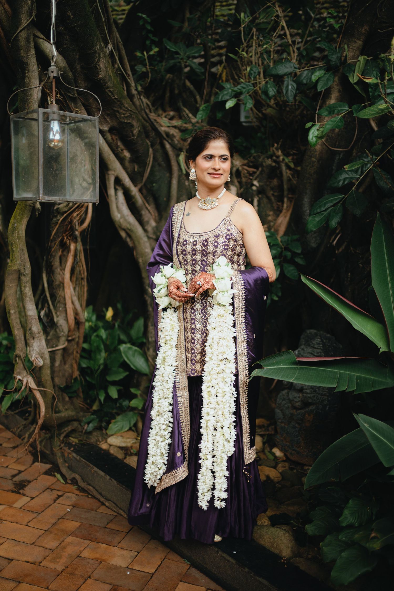 Indian bride in purple lehenga holding white floral garlands, posing outdoors among lush green trees.