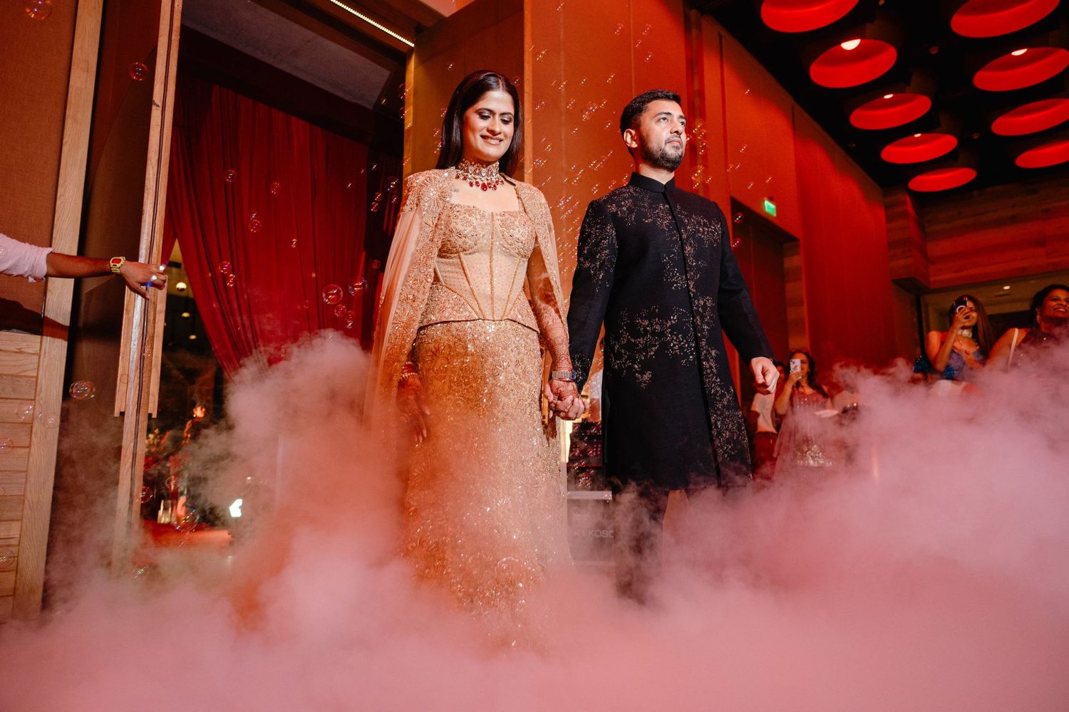 Couple in elegant Indian wedding attire walking through dramatic pink smoke during their grand reception entrance.