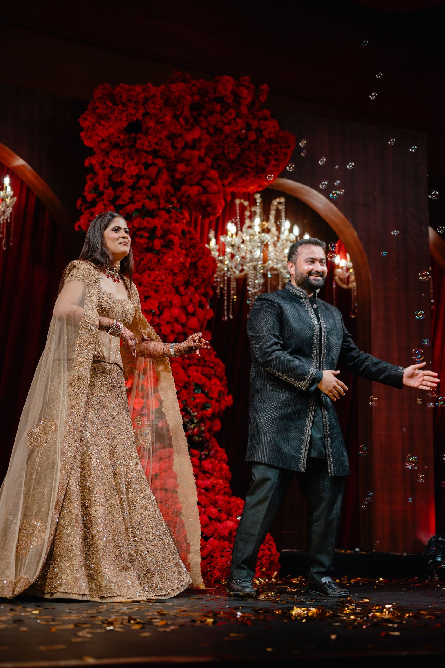 Couple in traditional Indian wedding attire stand on stage with red floral backdrop and chandelier, surrounded by bubbles.