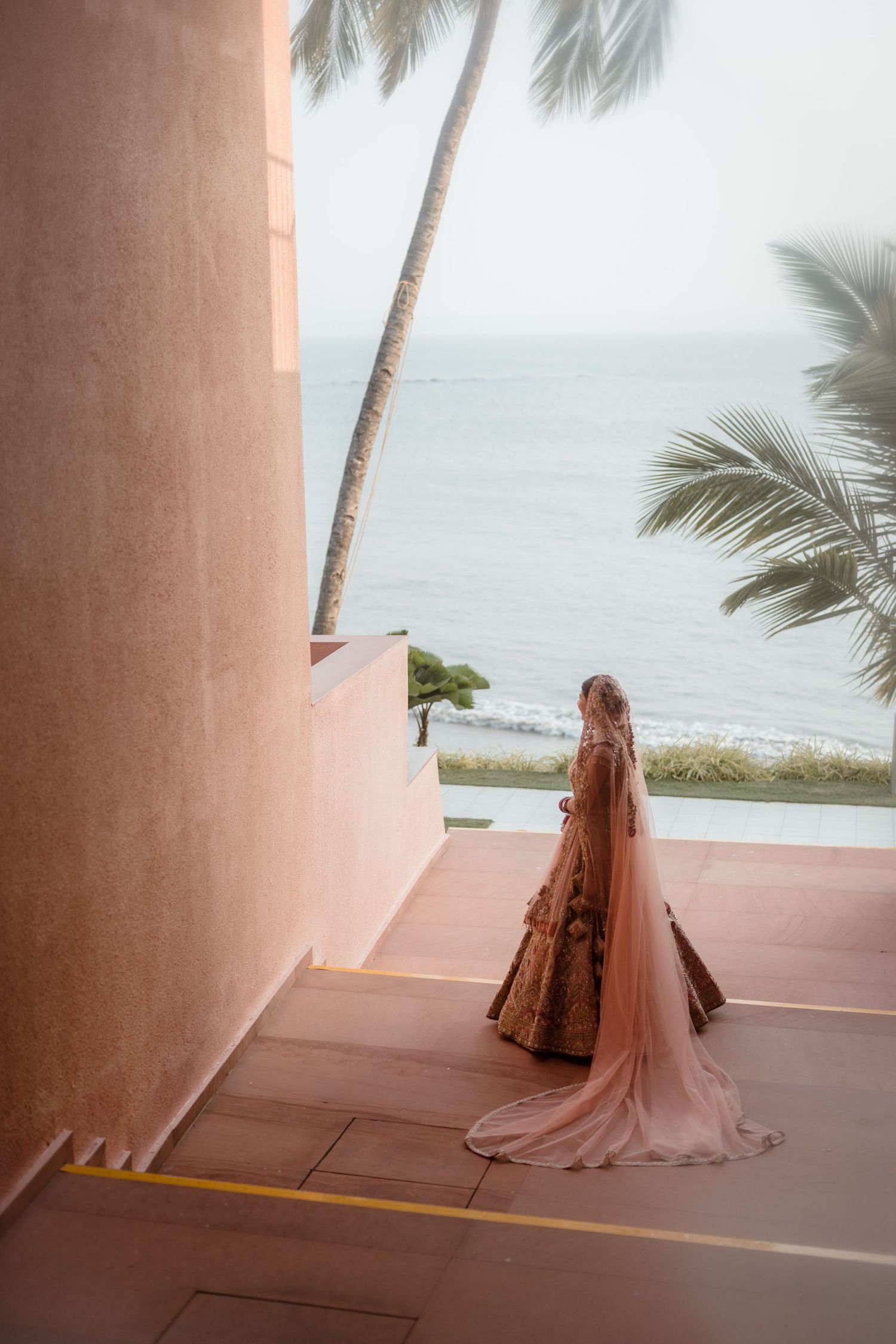 A person in a flowing gown stands on a pink balcony overlooking a tropical beach with palm trees and ocean views.