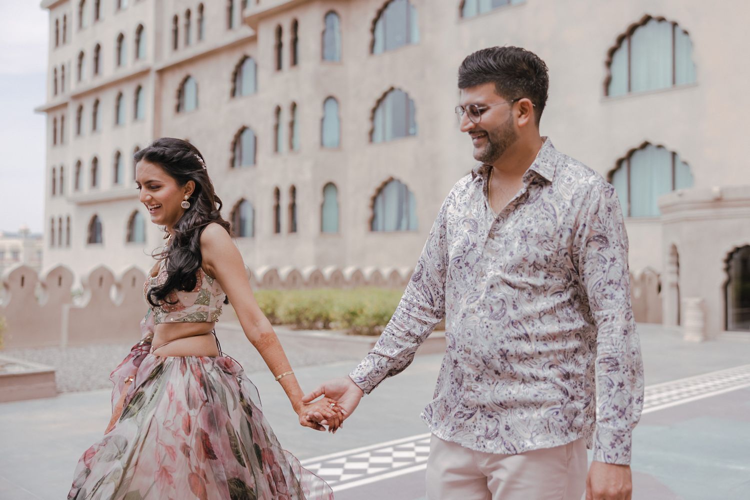 Couple in elegant formal attire holding hands while walking near a building with arched windows.