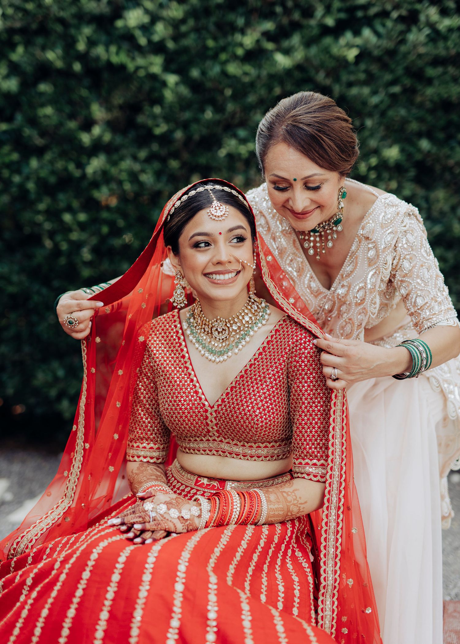 A bride in traditional red Indian lehenga shares a joyful moment while getting her veil adjusted in a garden setting.