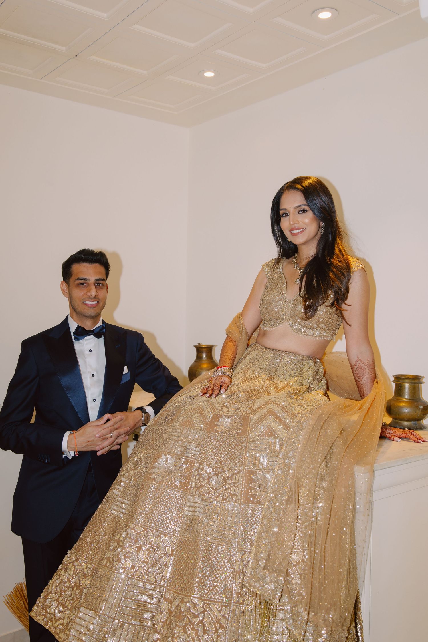 Couple in formal attire posing with gold sequined gown and tuxedo against white backdrop.