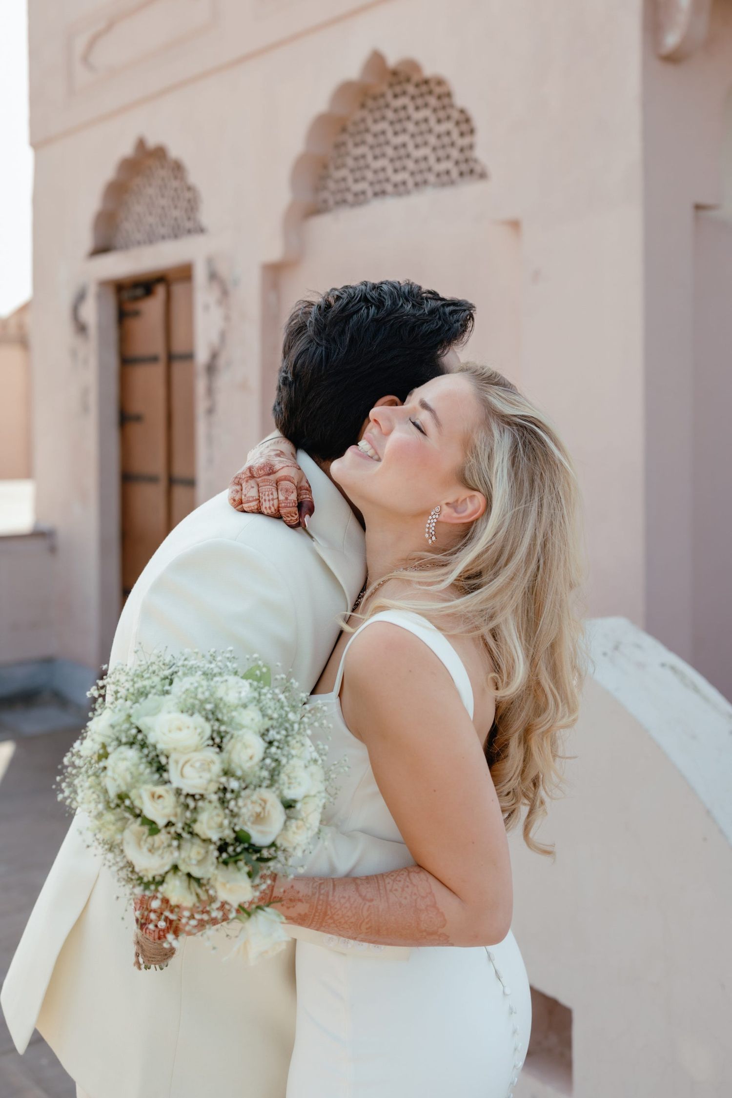 Romantic moment captured between couple embracing with white bouquet against pink architectural backdrop.