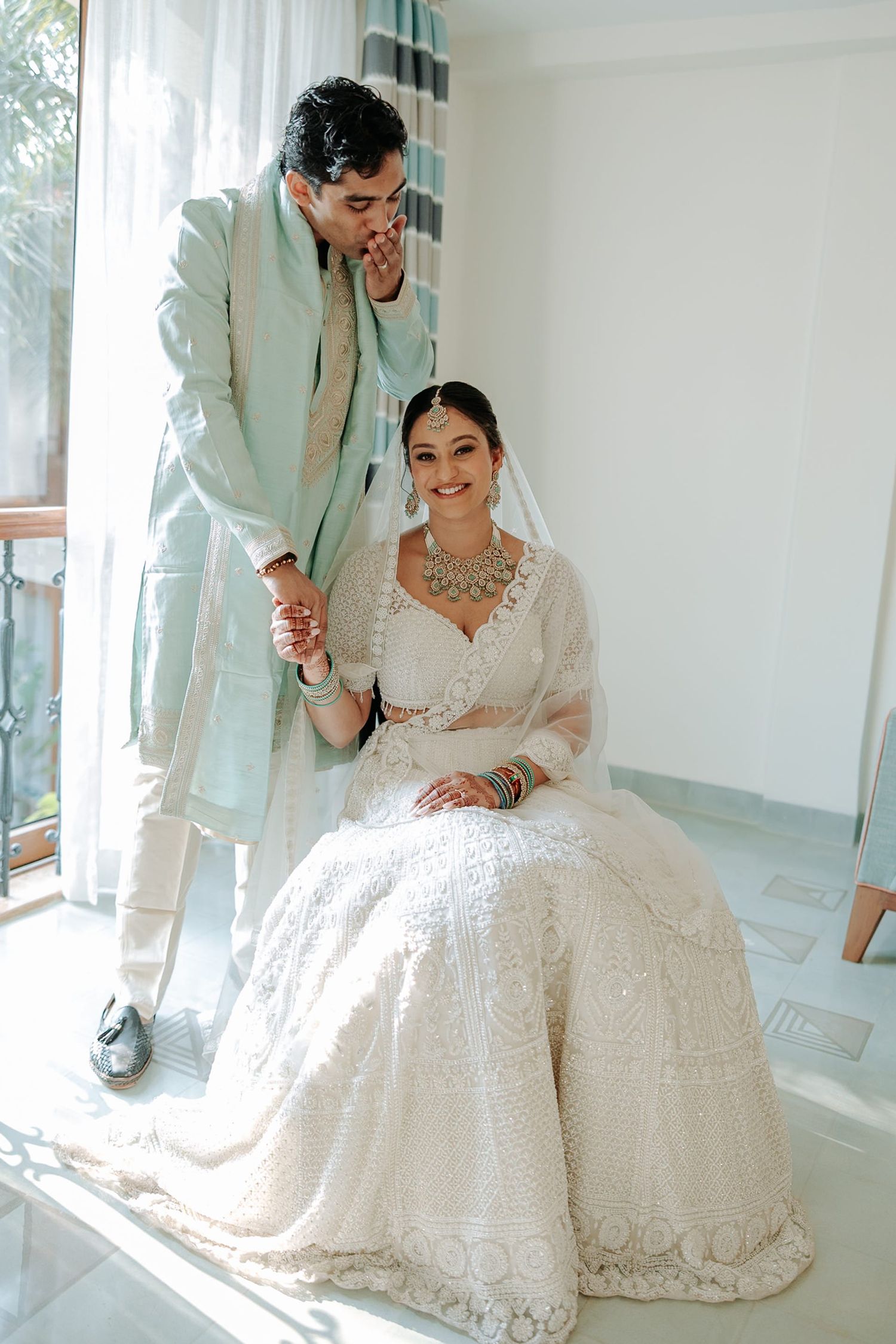 Indian bride in white lehenga seated with groom in mint sherwani holding hands in elegant bridal portrait.