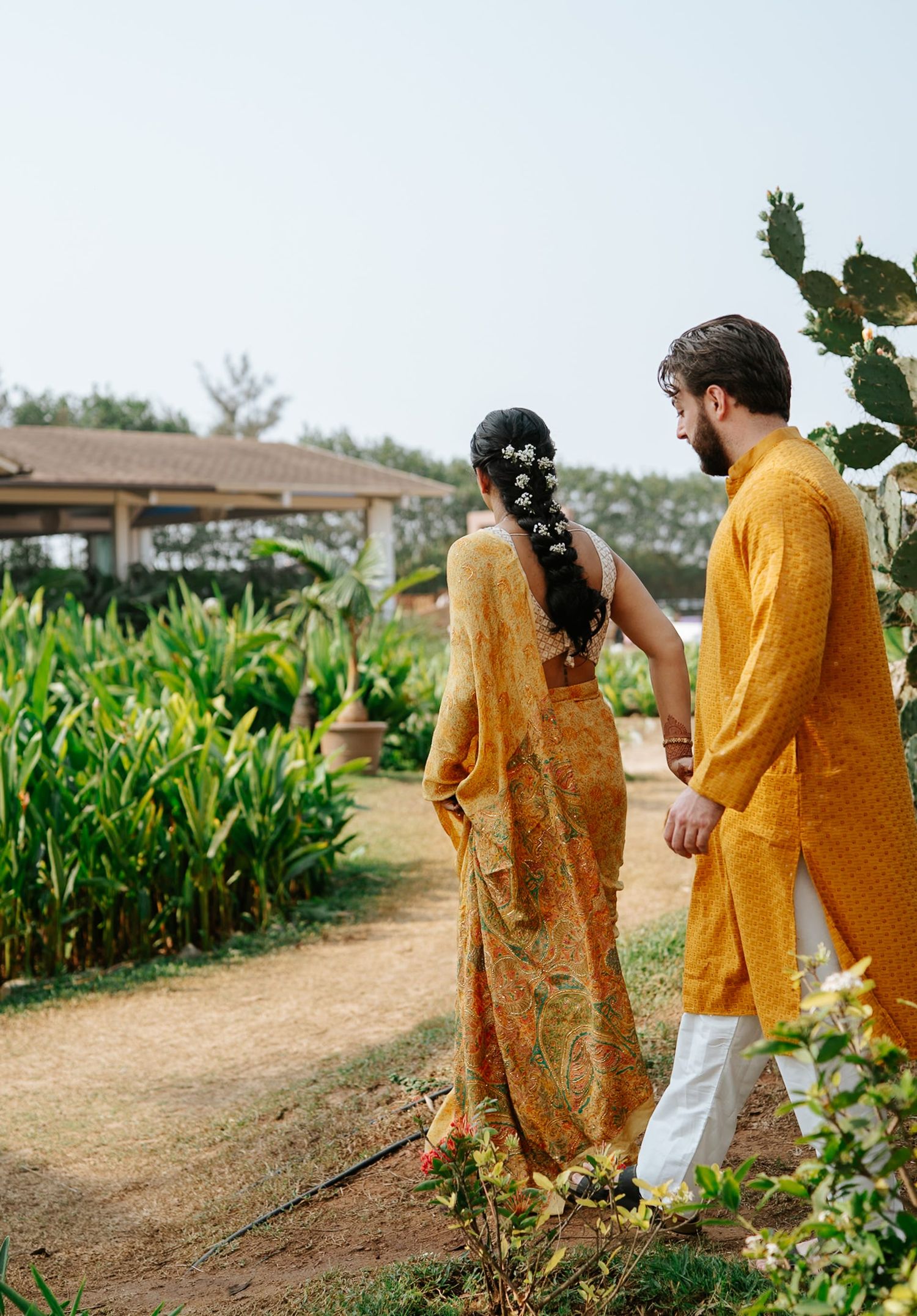 Indian couple in matching yellow outfits walking hand in hand through a lush garden path on their wedding day.