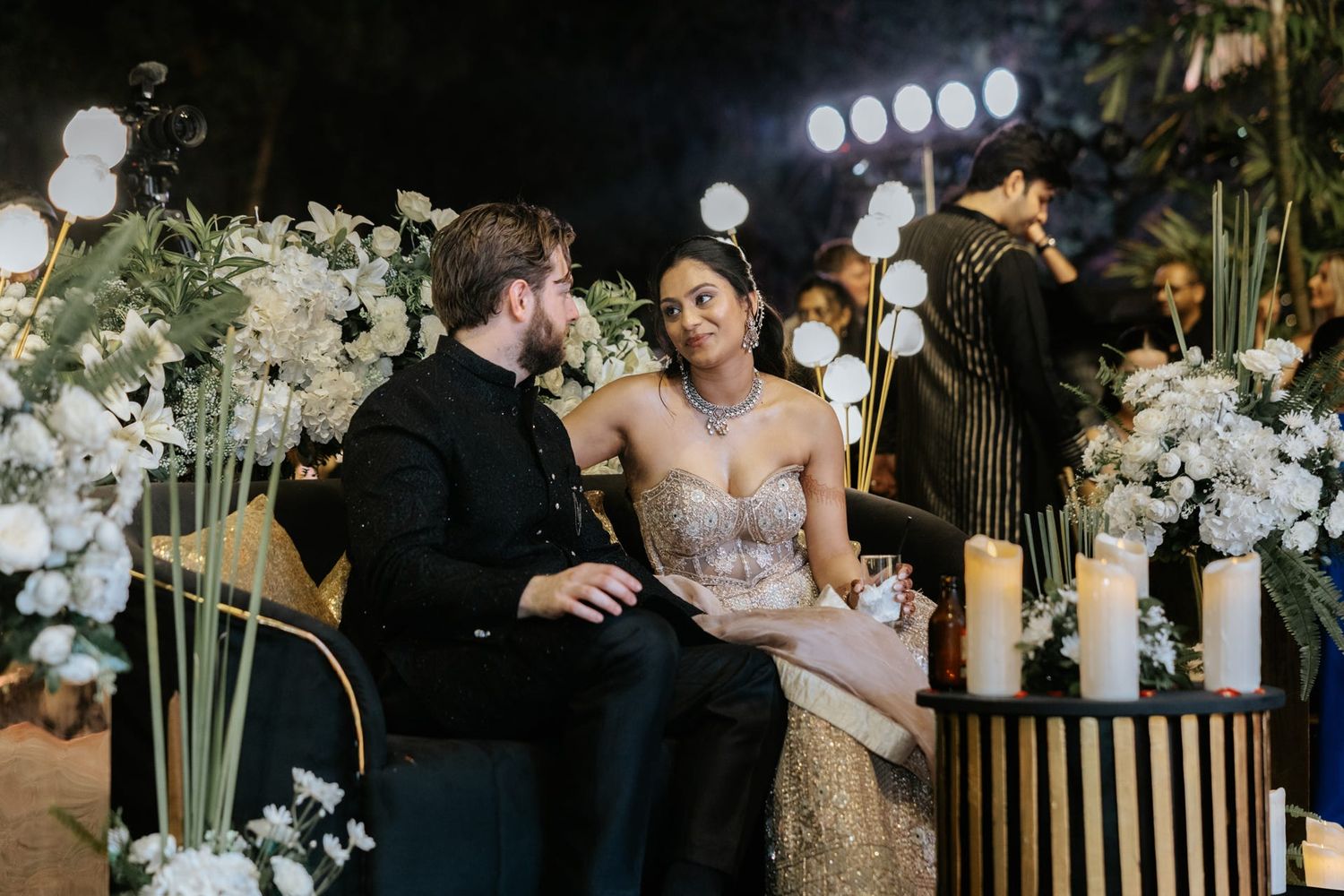 Couple in elegant attire seated together at a romantic wedding reception with white floral arrangements and candles.