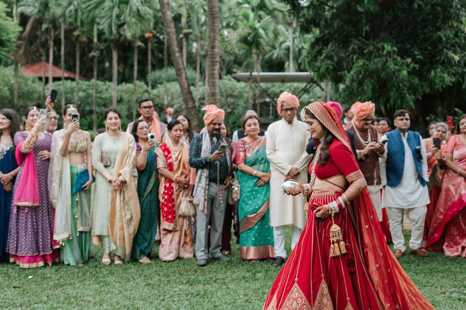 Indian wedding guests in colorful traditional attire watch the bride in a red lehenga during an outdoor ceremony.