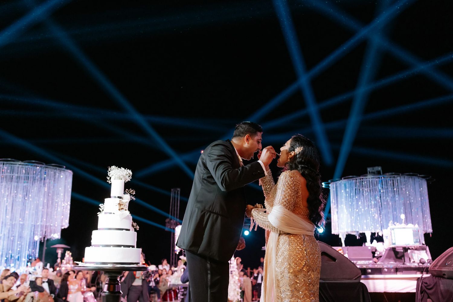 Couple sharing wedding cake at elegant reception with blue laser lights and white tiered cake on display.