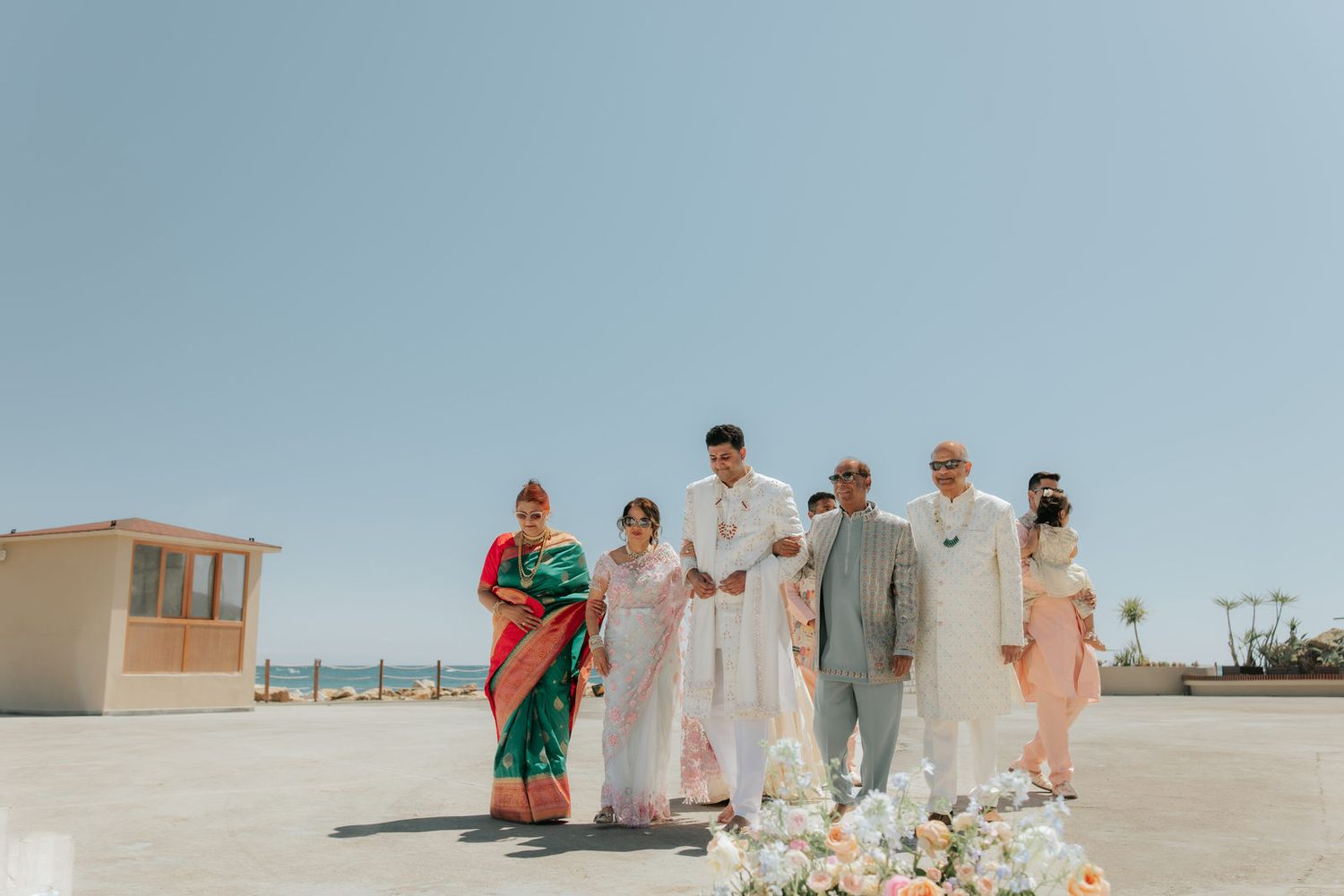 Indian wedding group photo outdoors near beach, guests in colorful traditional attire including sarees, under clear blue sky.