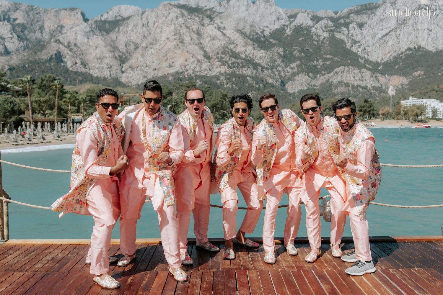 Seven groomsmen in matching pink suits pose on a waterfront deck with mountain views.