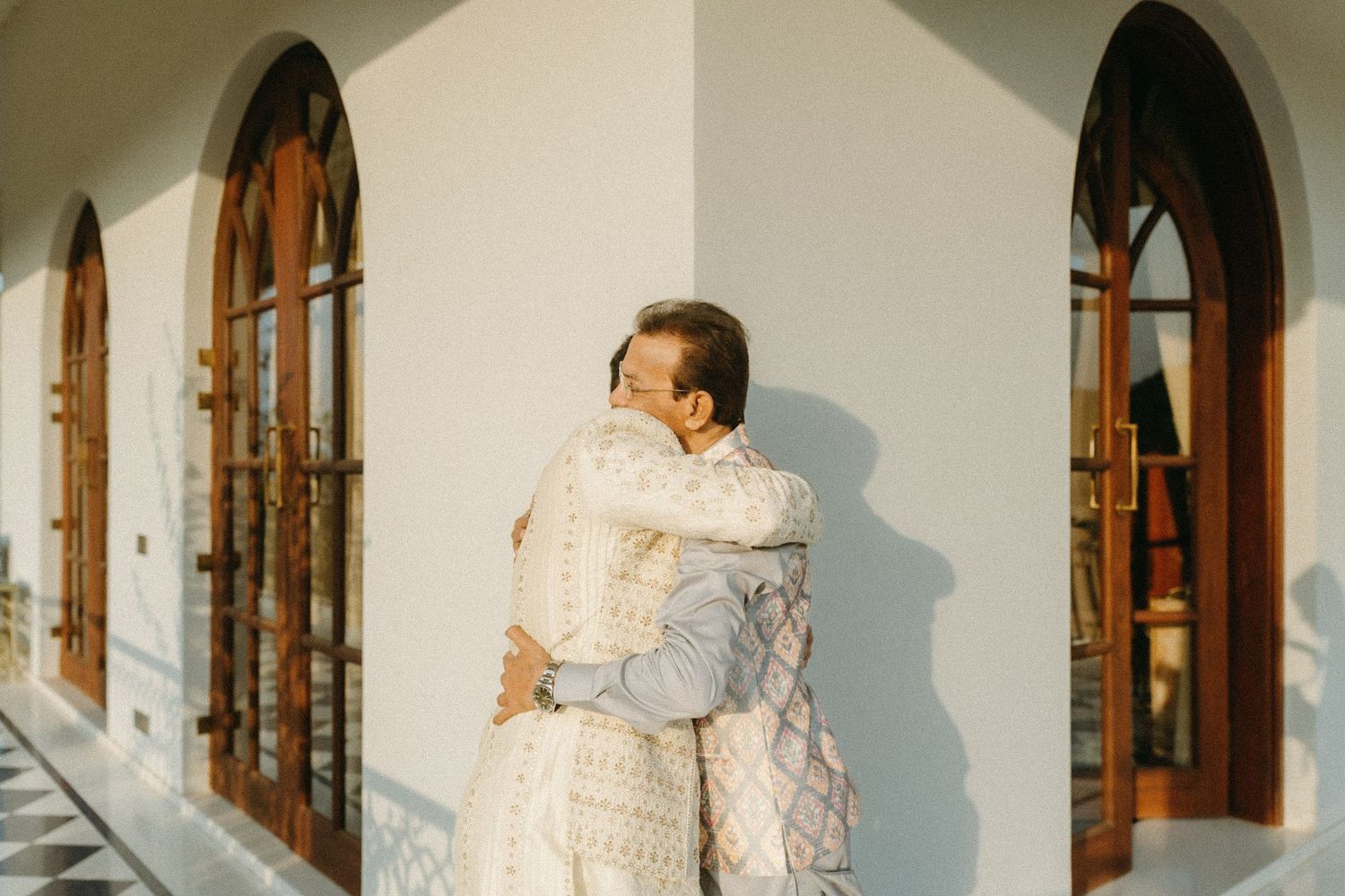 A couple in elegant white outfits embrace against a white wall with arched windows casting warm sunlit shadows.