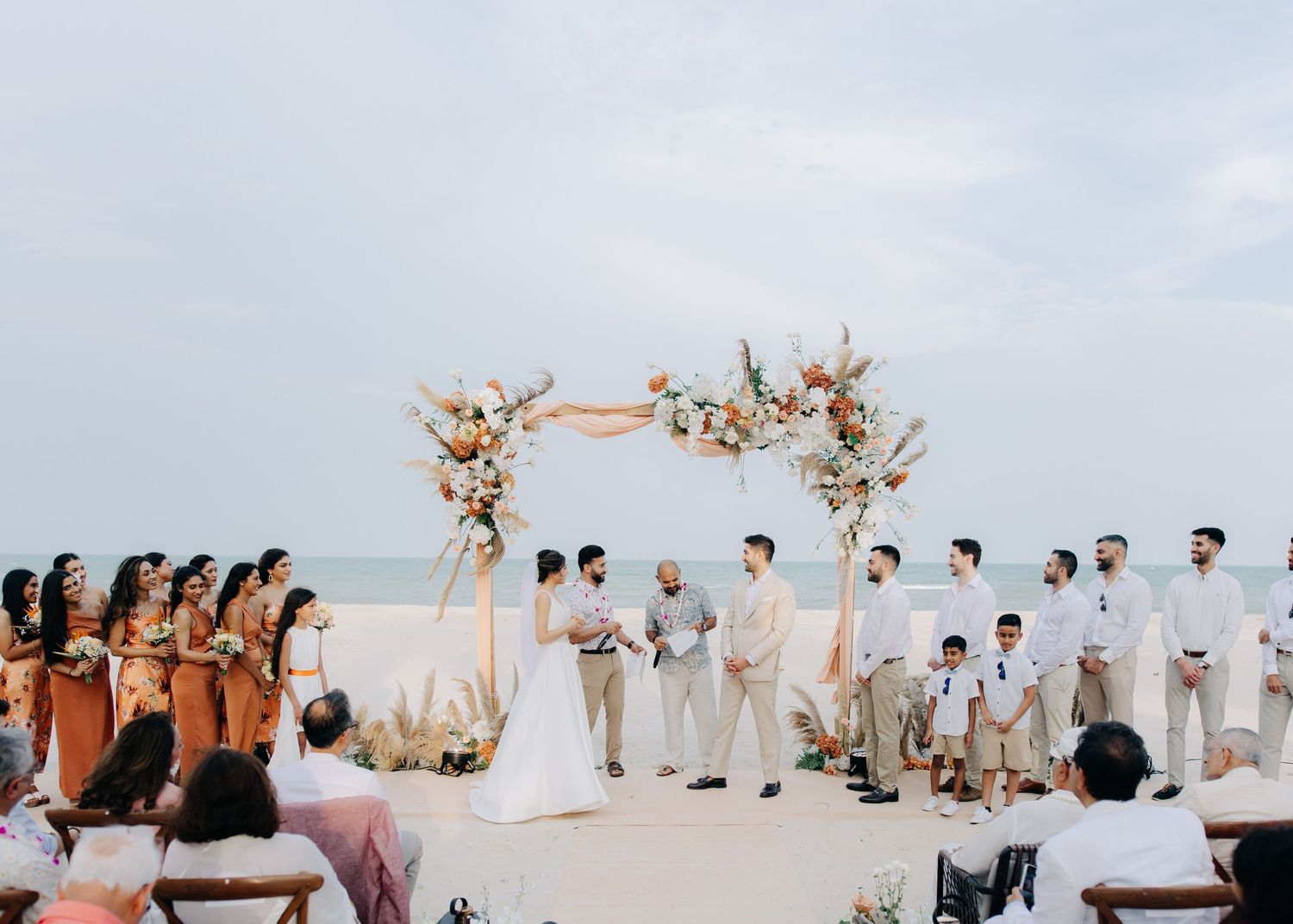 A beach wedding ceremony with bridesmaids in rust-colored dresses, groomsmen in white, under a floral arch at sunset.