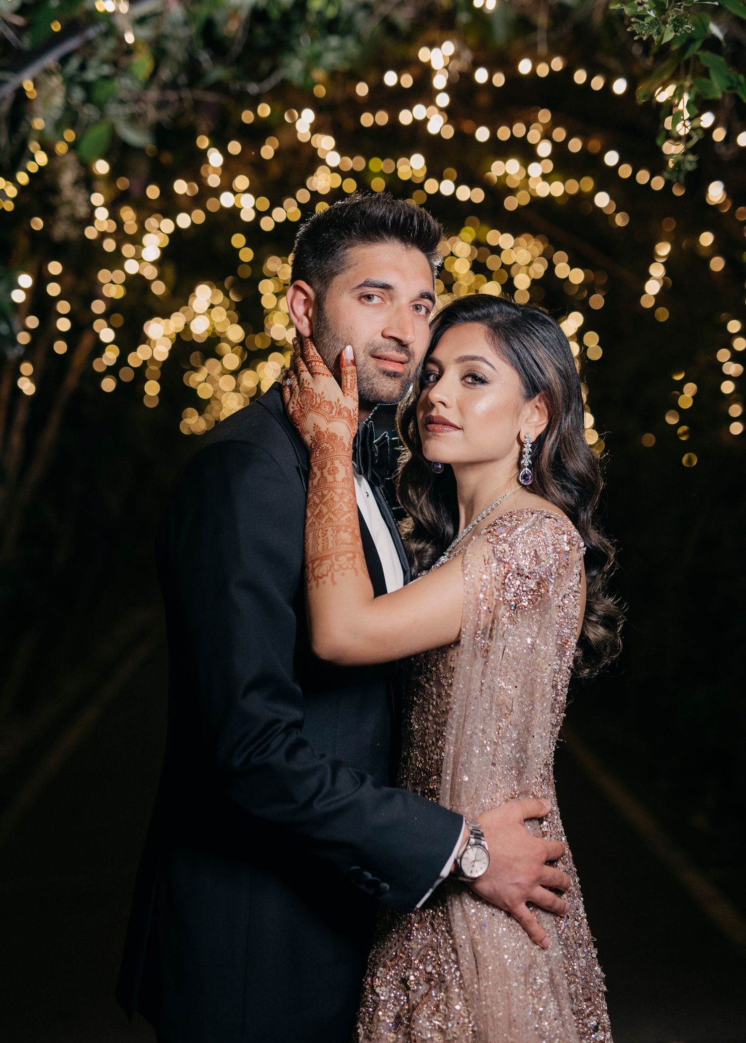 Romantic couple embraces under twinkling fairy lights at night, with woman wearing sparkly rose gold dress and man in dark suit.