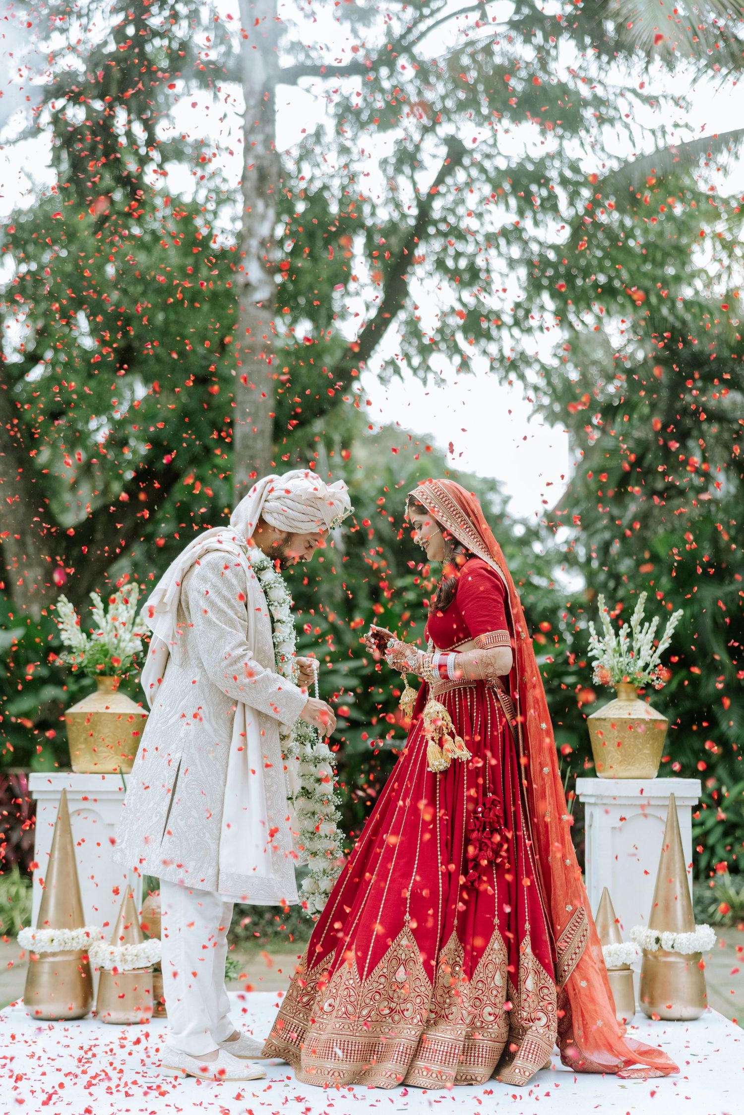 Indian bride in red lehenga and groom in white sherwani exchange vows outdoors with flower petals falling.
