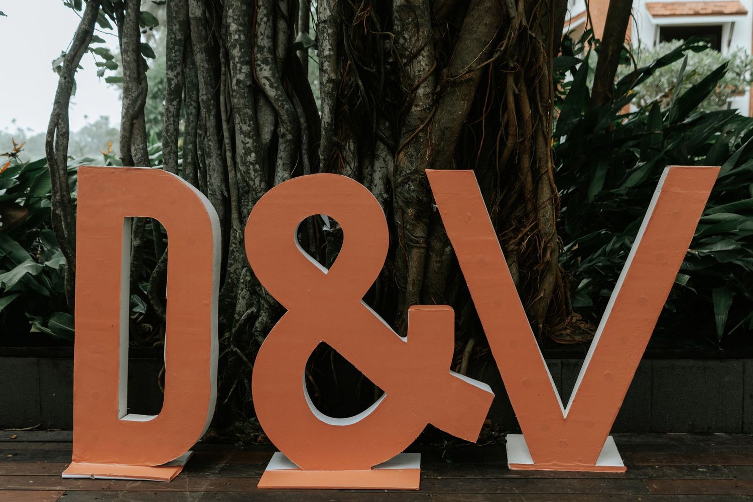 Large terracotta letter signs spelling D&V displayed outdoors against lush tropical trees at a wedding venue.