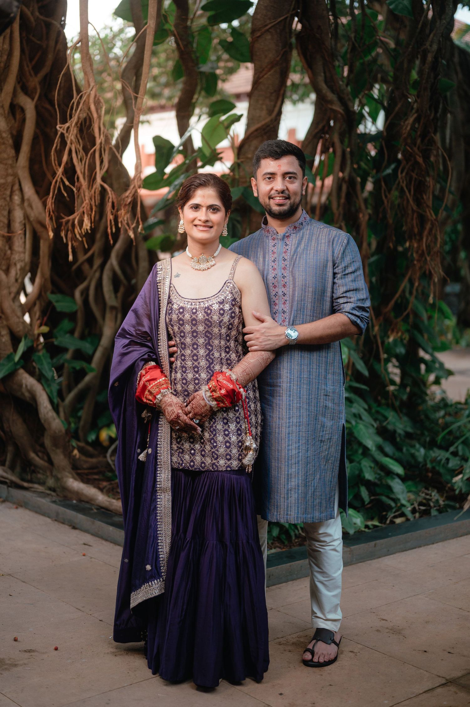 Indian couple in traditional attire, woman in navy sharara suit with dupatta, man in grey kurta, posing outdoors.