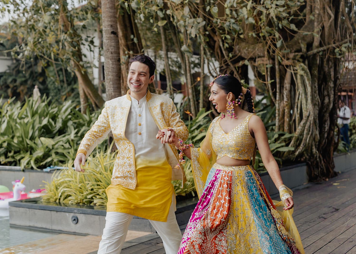 Couple walking together outdoors wearing coordinated yellow and multicolored Indian wedding outfits.