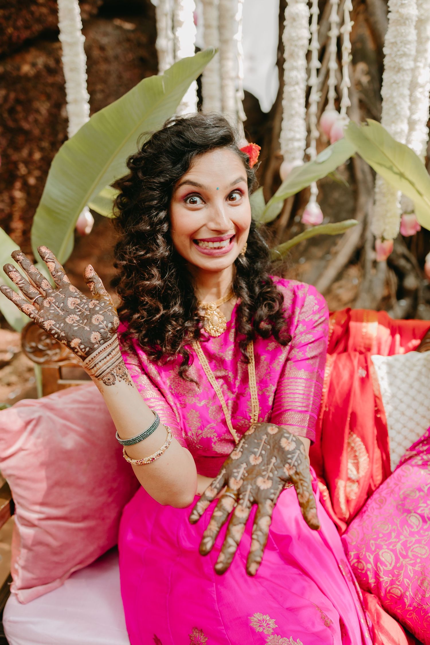 Bride in pink saree shows intricate mehndi henna designs on hands, smiling joyfully at outdoor wedding celebration.