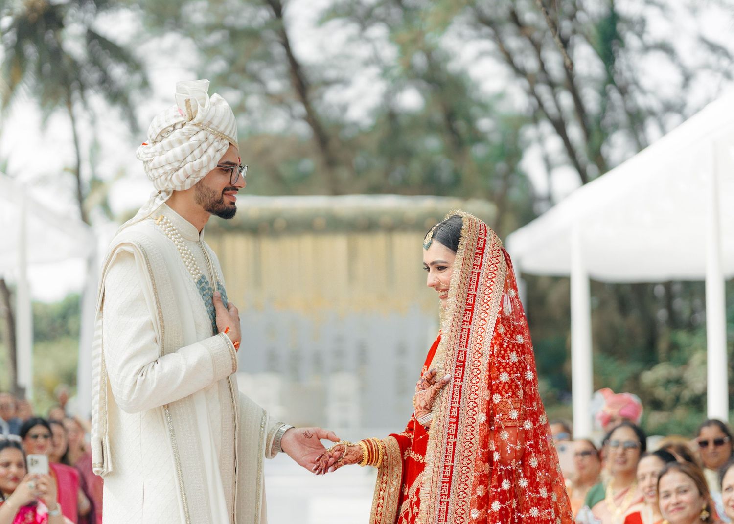 A couple in traditional Indian wedding attire exchanges vows during an outdoor ceremony with guests looking on.