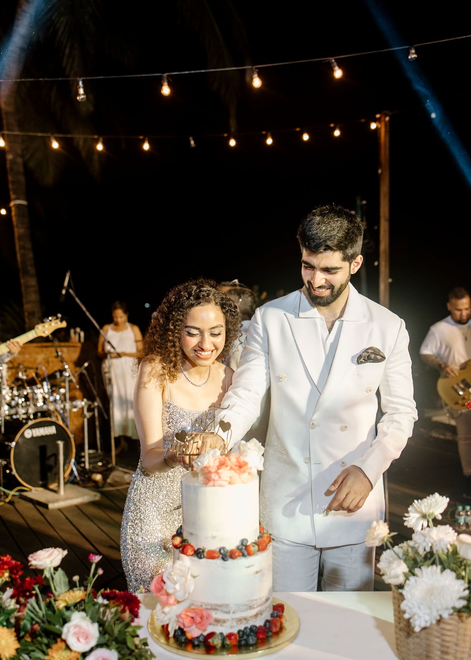 Wedding cake cutting celebration with string lights and floral decorations at night.