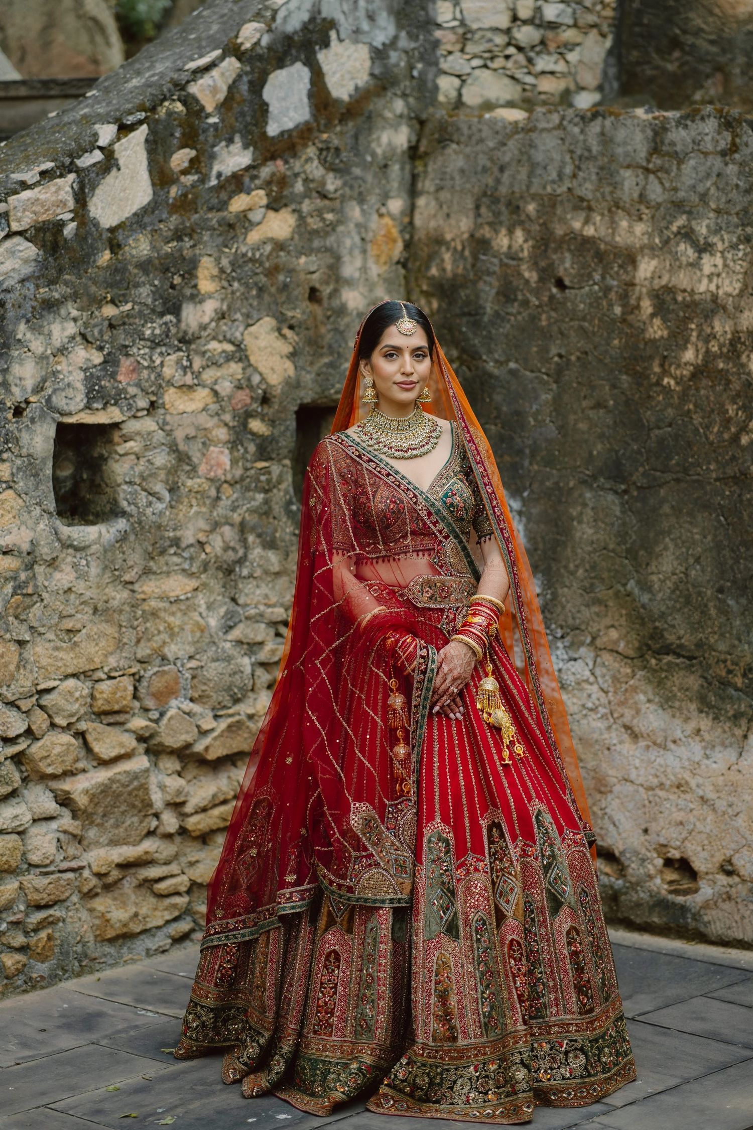 A bride in an ornate red lehenga poses against ancient stone walls.