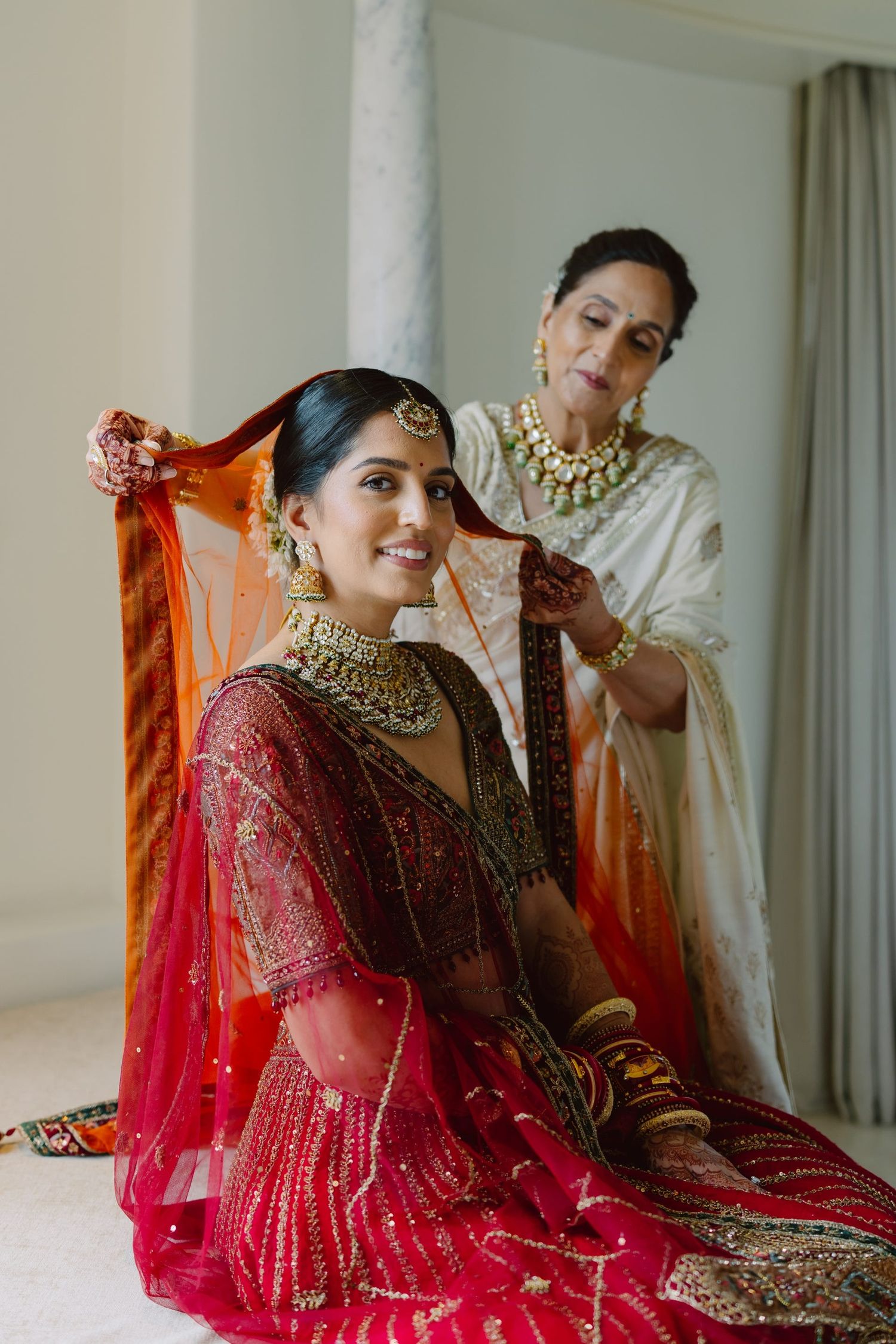 A bride in red lehenga getting her dupatta adjusted during wedding preparations.