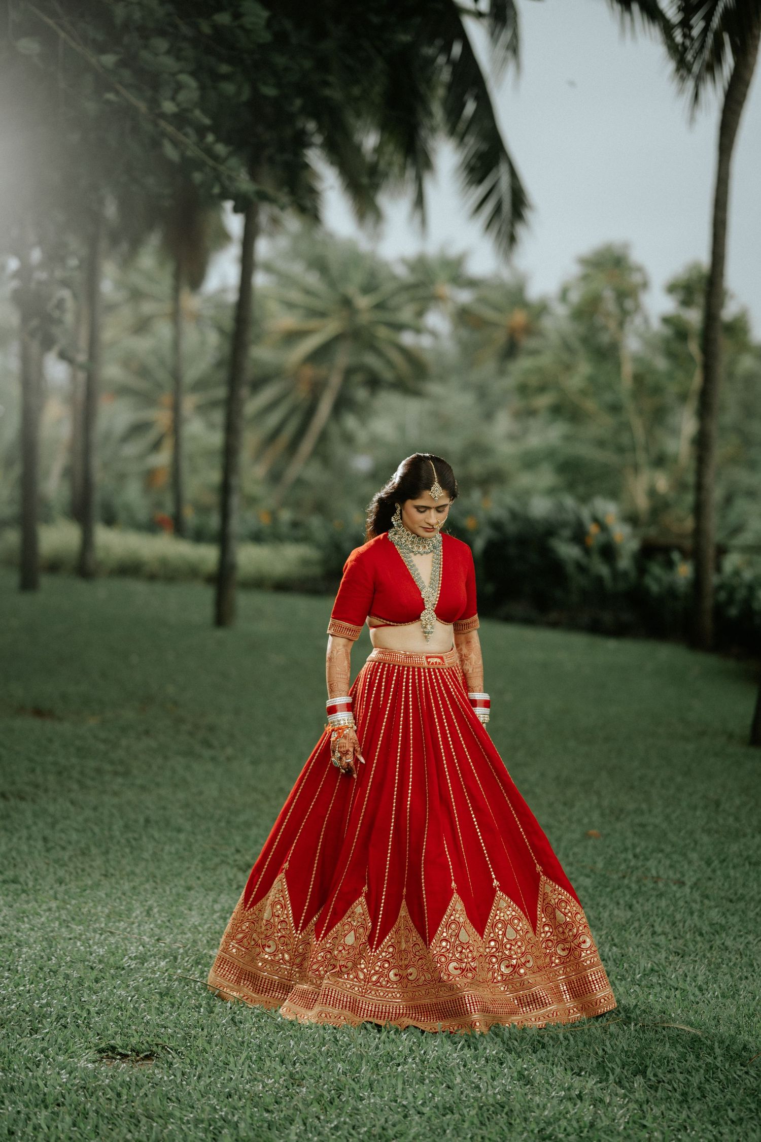 Indian bride in stunning red and gold lehenga standing gracefully in a lush green garden with palm trees.
