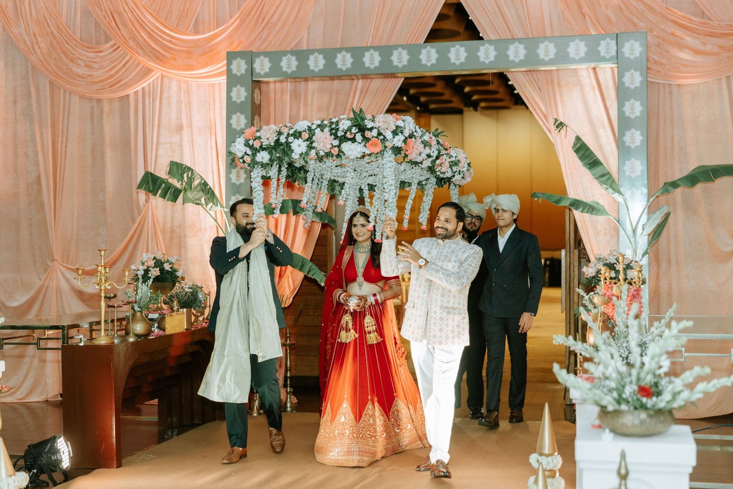 Bride in red lehenga and groom in traditional Indian wedding attire make grand entrance at decorated venue.