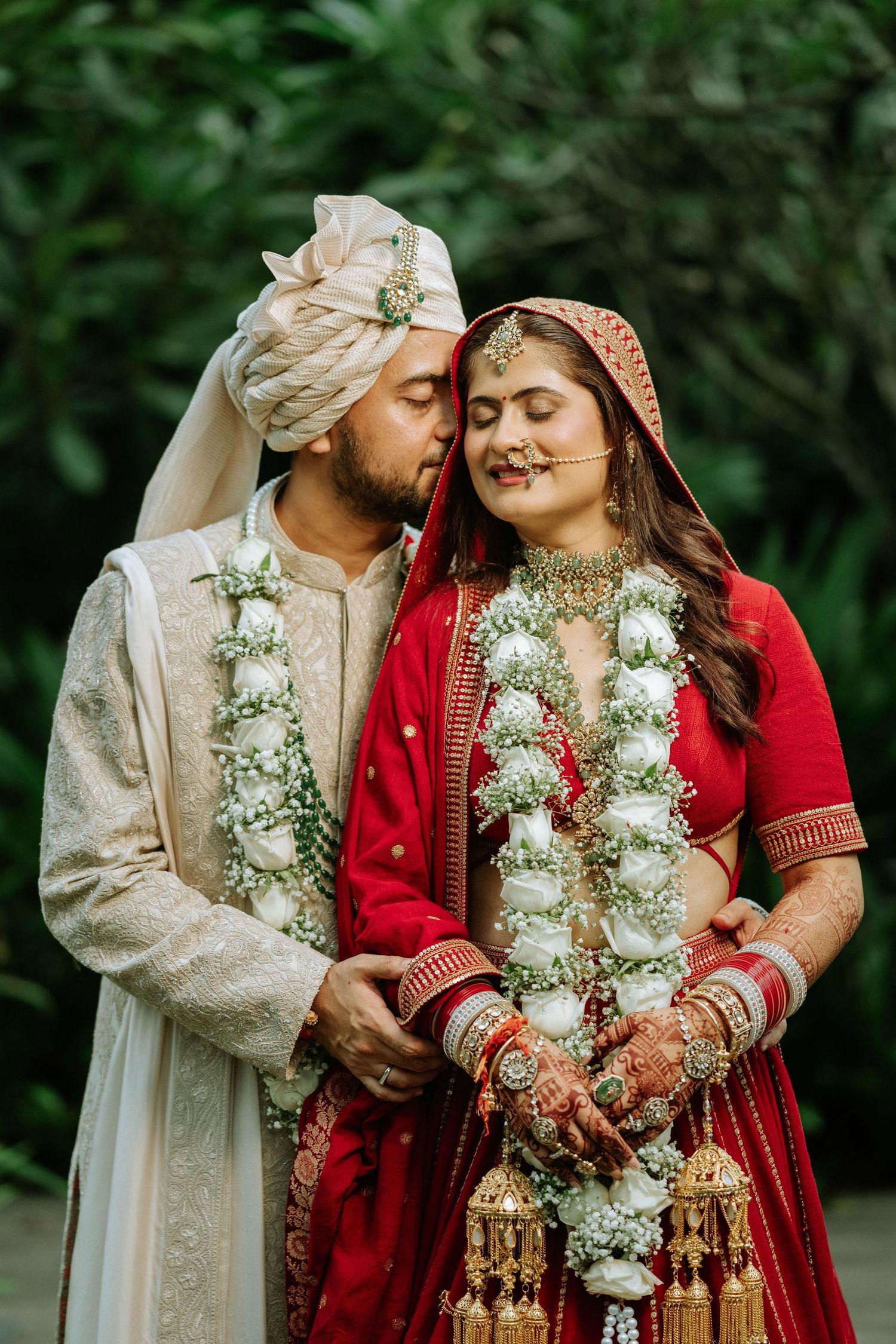 Indian couple in traditional wedding attire, groom in cream sherwani kissing bride in red lehenga with floral garlands.