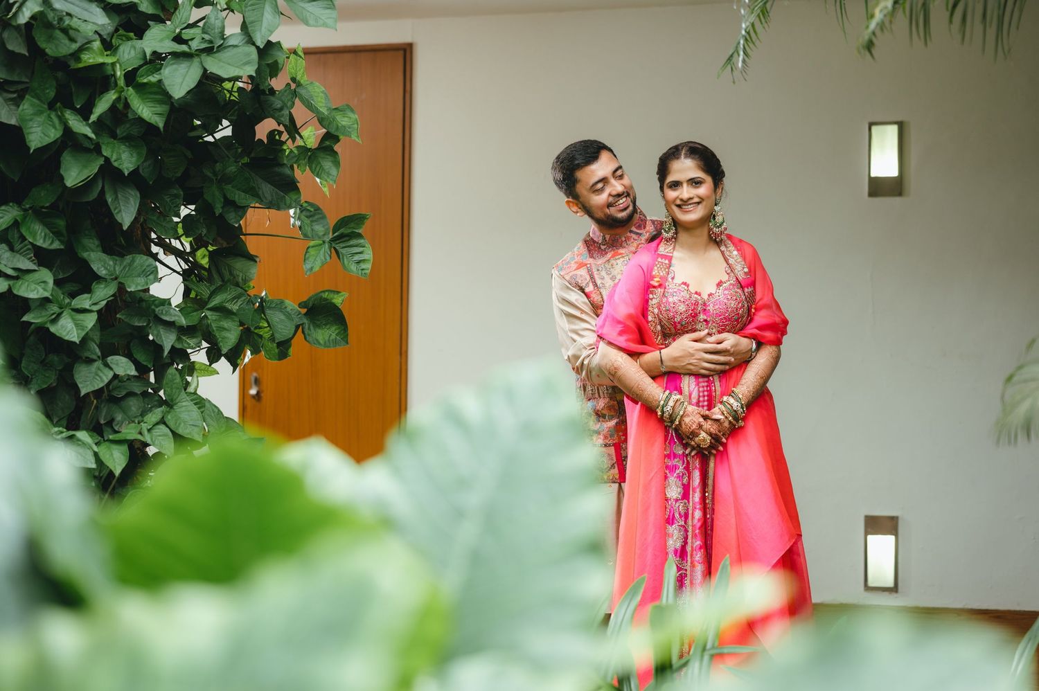 Couple in matching pink traditional Indian outfits smiling together outdoors among lush green tropical plants.