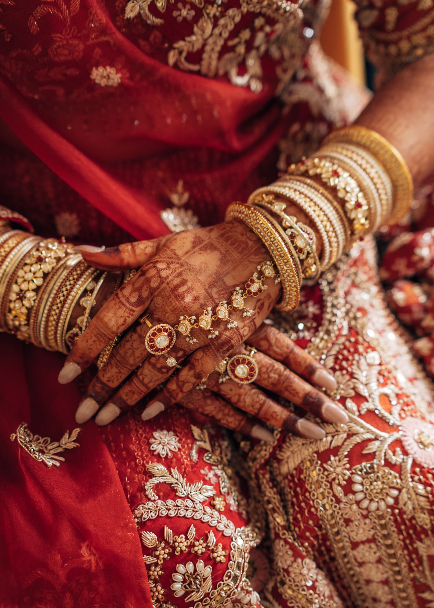 Close-up of Indian bride's hands adorned with intricate henna, gold bangles, and rings against a red embroidered sari.